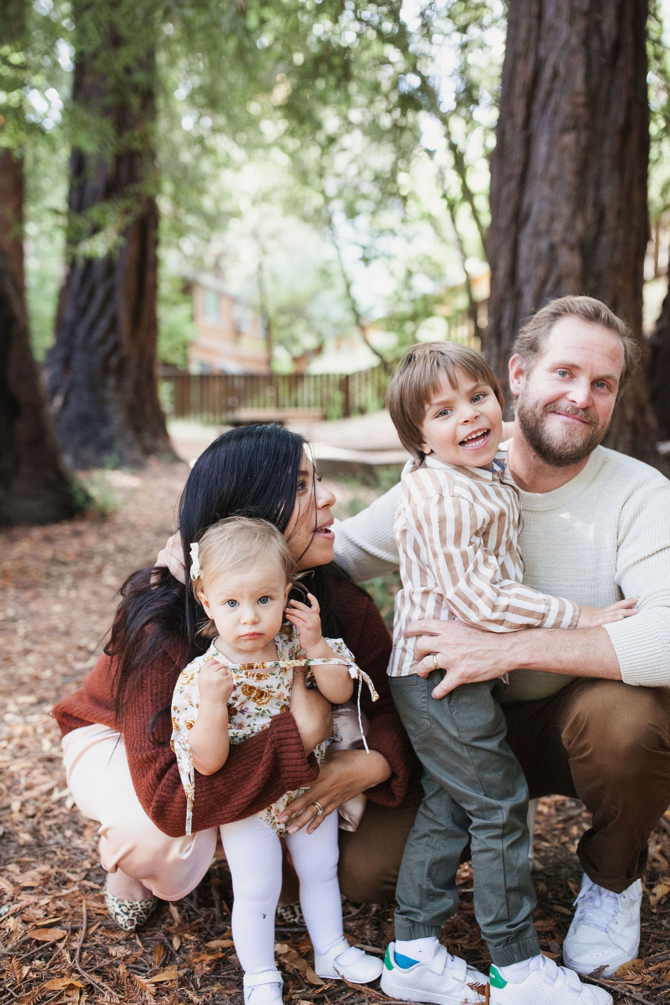 A family of four crouches together among towering redwoods, their genuine smiles catching dappled sunlight filtering through the canopy