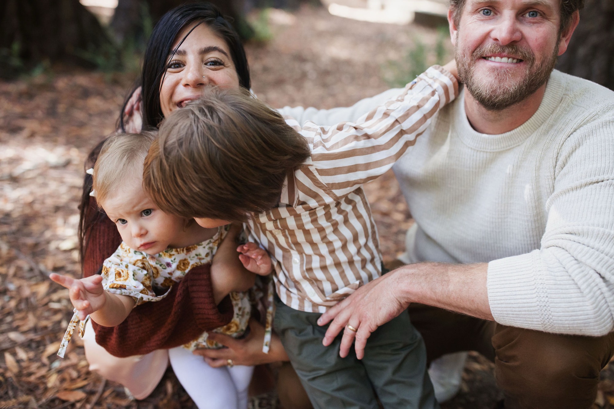 A bearded man and dark-haired woman crouch beside two young children on a forest floor, all smiling warmly at the camera amid dappled woodland light