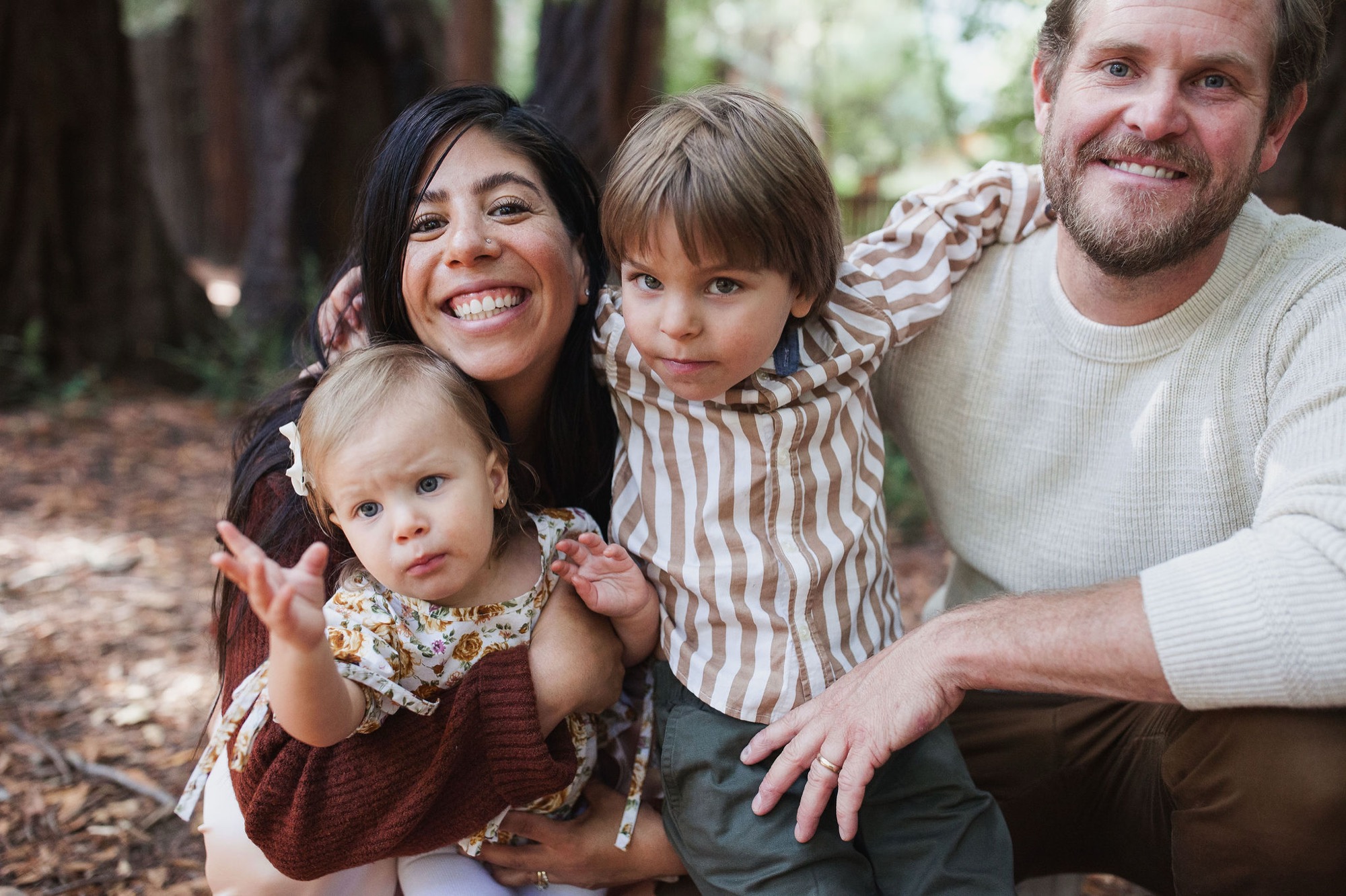 A smiling family of four sits together on forest ground, with two young children nestled between their parents amid autumn leaves and natural woodland surroundings