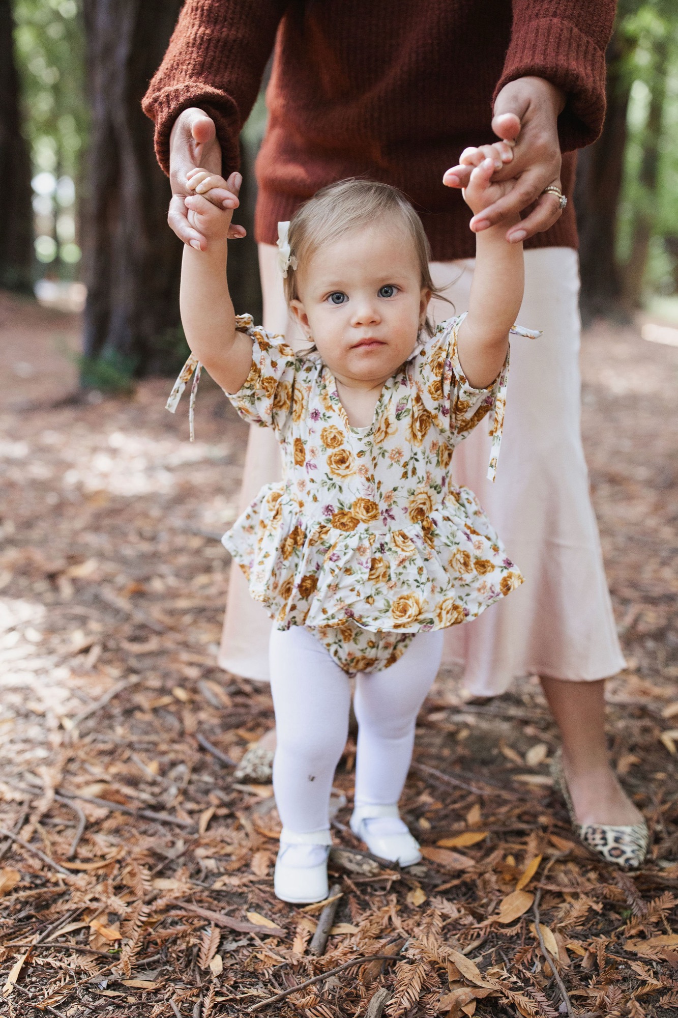 A toddler in a mustard floral dress holds both hands of an adult in a rust sweater while standing on a leafy forest path