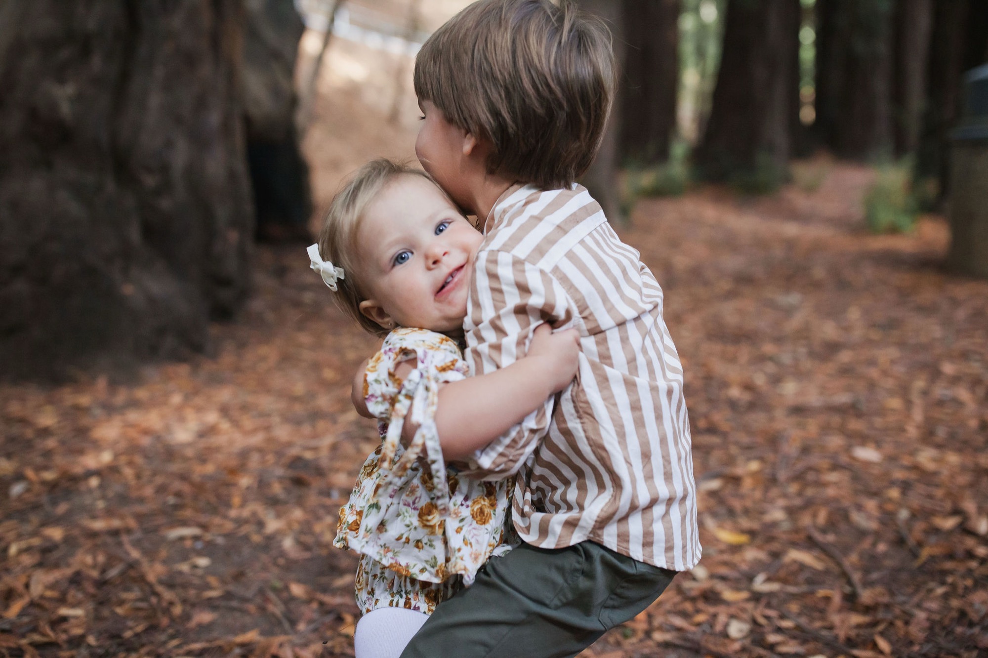 A boy in a striped shirt holds his younger sister close on a forest trail, both gazing toward the camera with gentle expressions