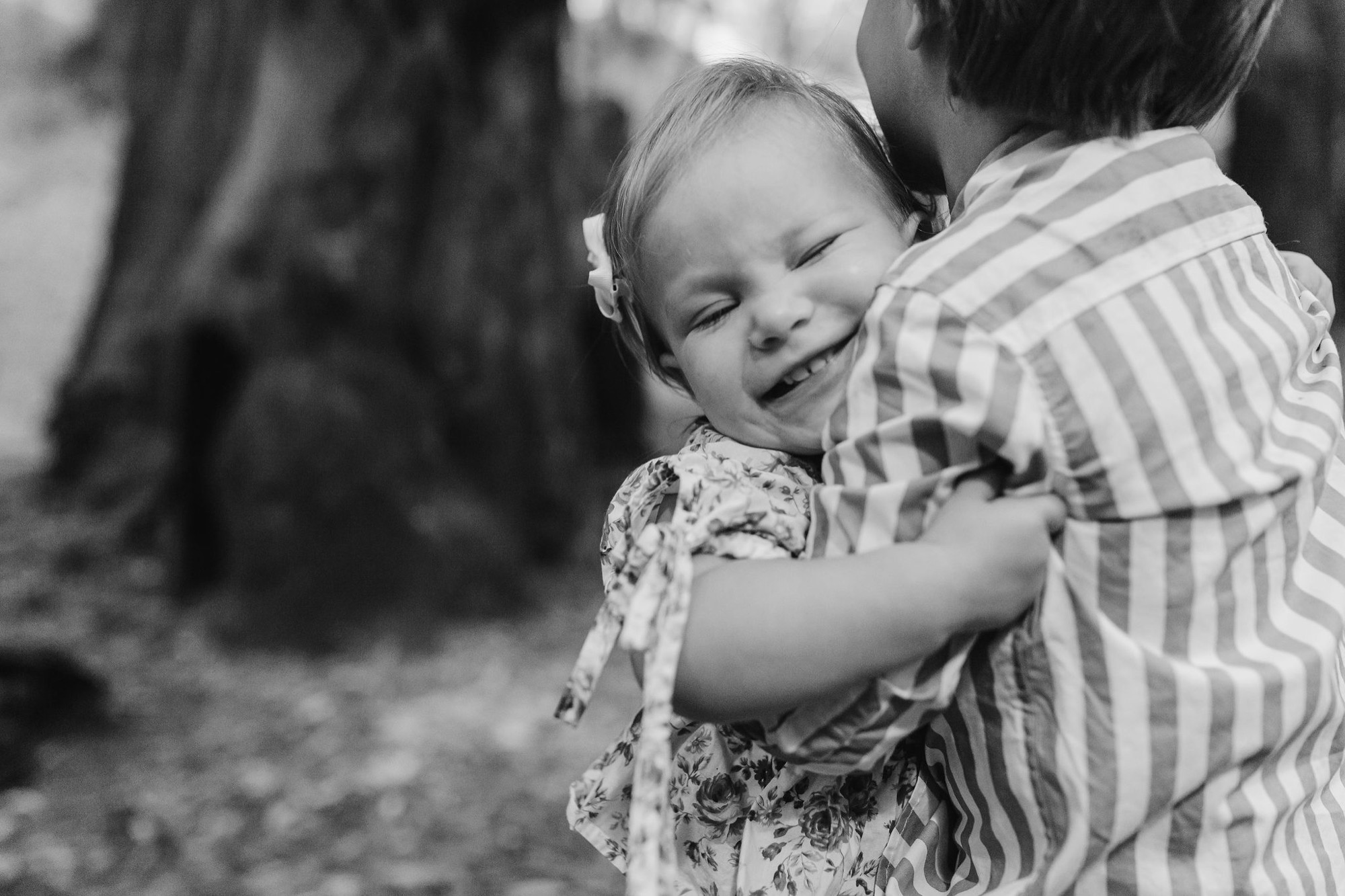 A young child with eyes closed smiles broadly while being held and embraced by an adult in a striped shirt, captured in soft black and white light