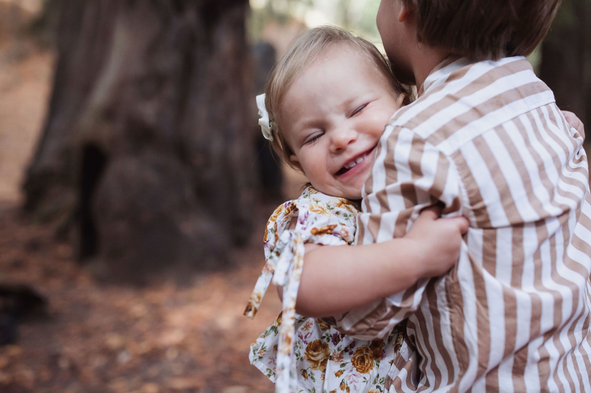 A toddler in a floral dress beams with joy while being held and hugged by an adult in a striped shirt