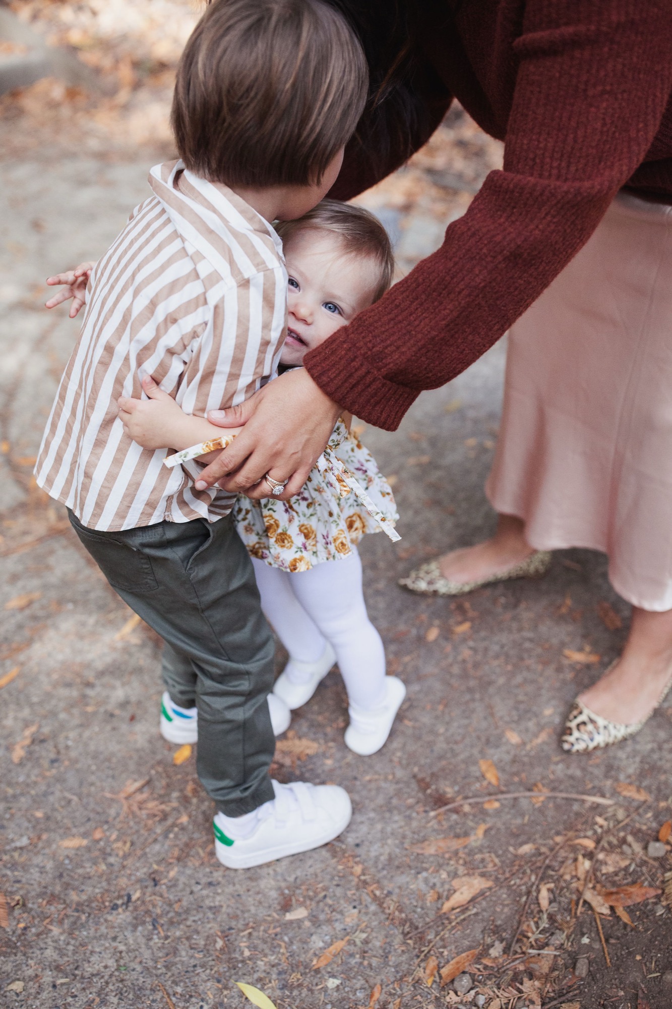 A toddler in striped shirt and white socks stands between two adults on pavement, holding hands in warm afternoon light
