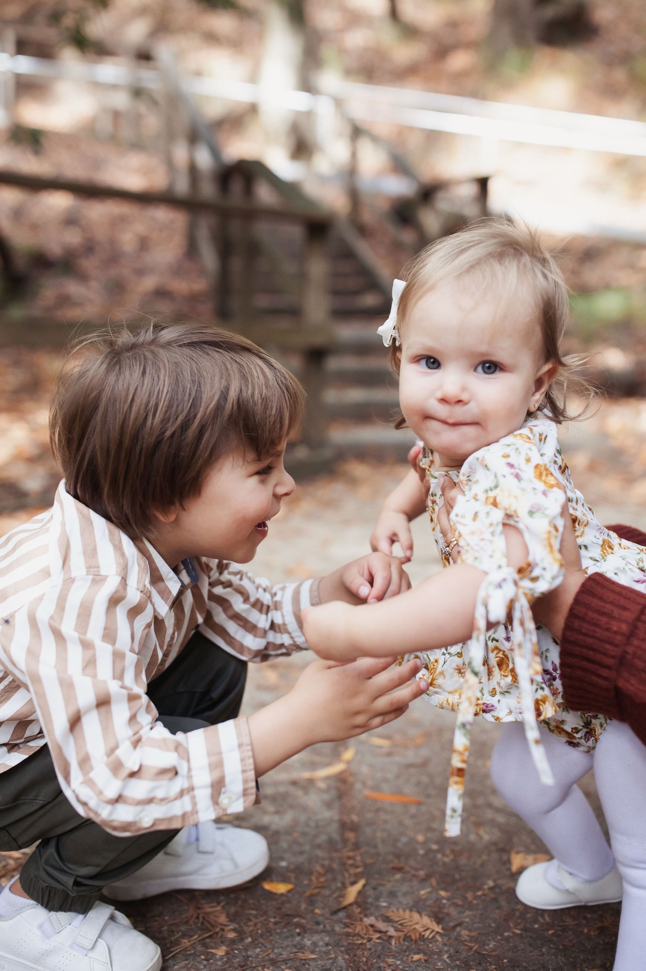 A young boy in a striped shirt gently holds hands with a toddler girl in a floral dress, both gazing downward with soft expressions on a dirt path