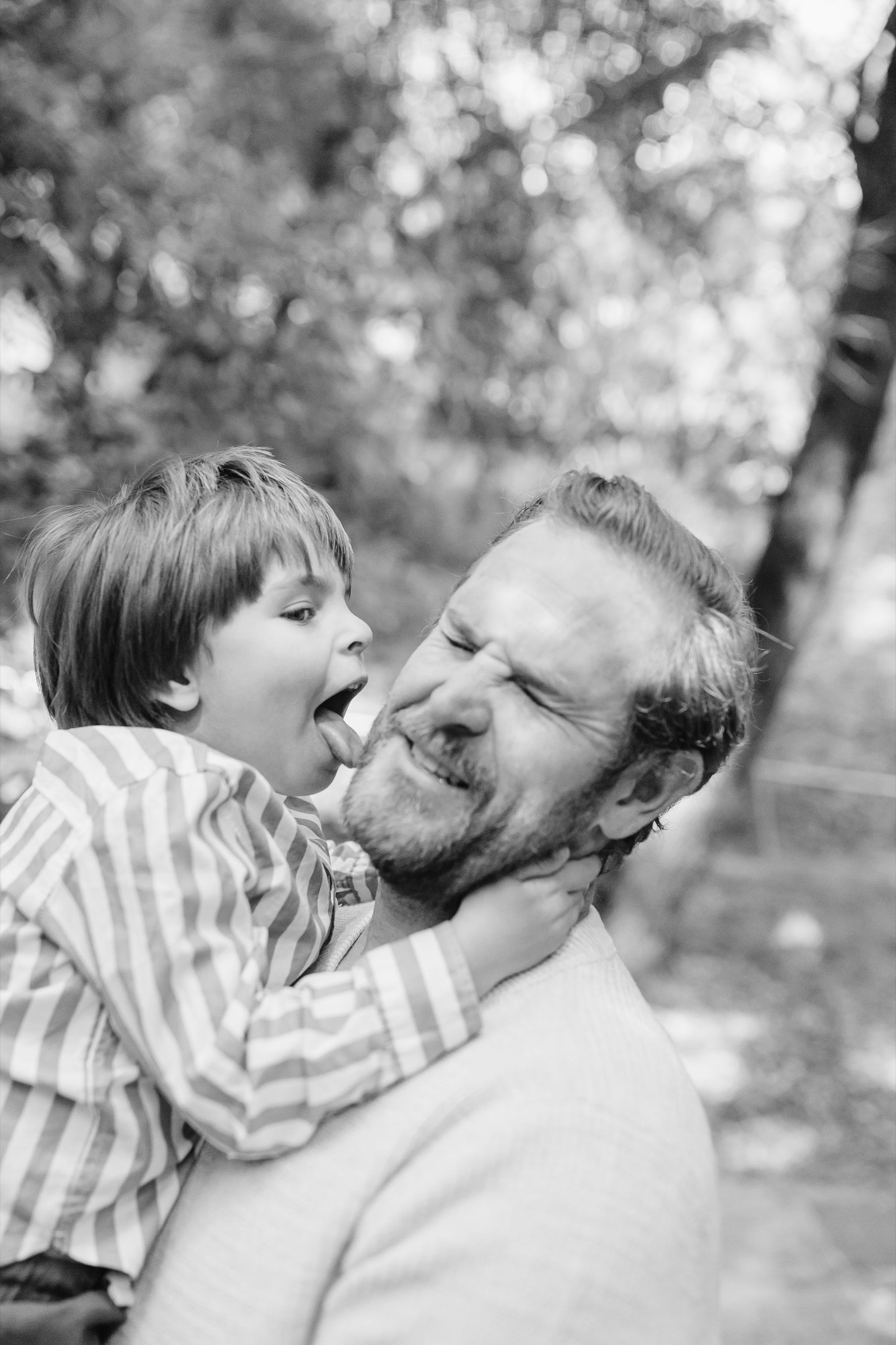 A man holds his young son at shoulder height in a striped shirt, both laughing with genuine delight against a soft-focused leafy background