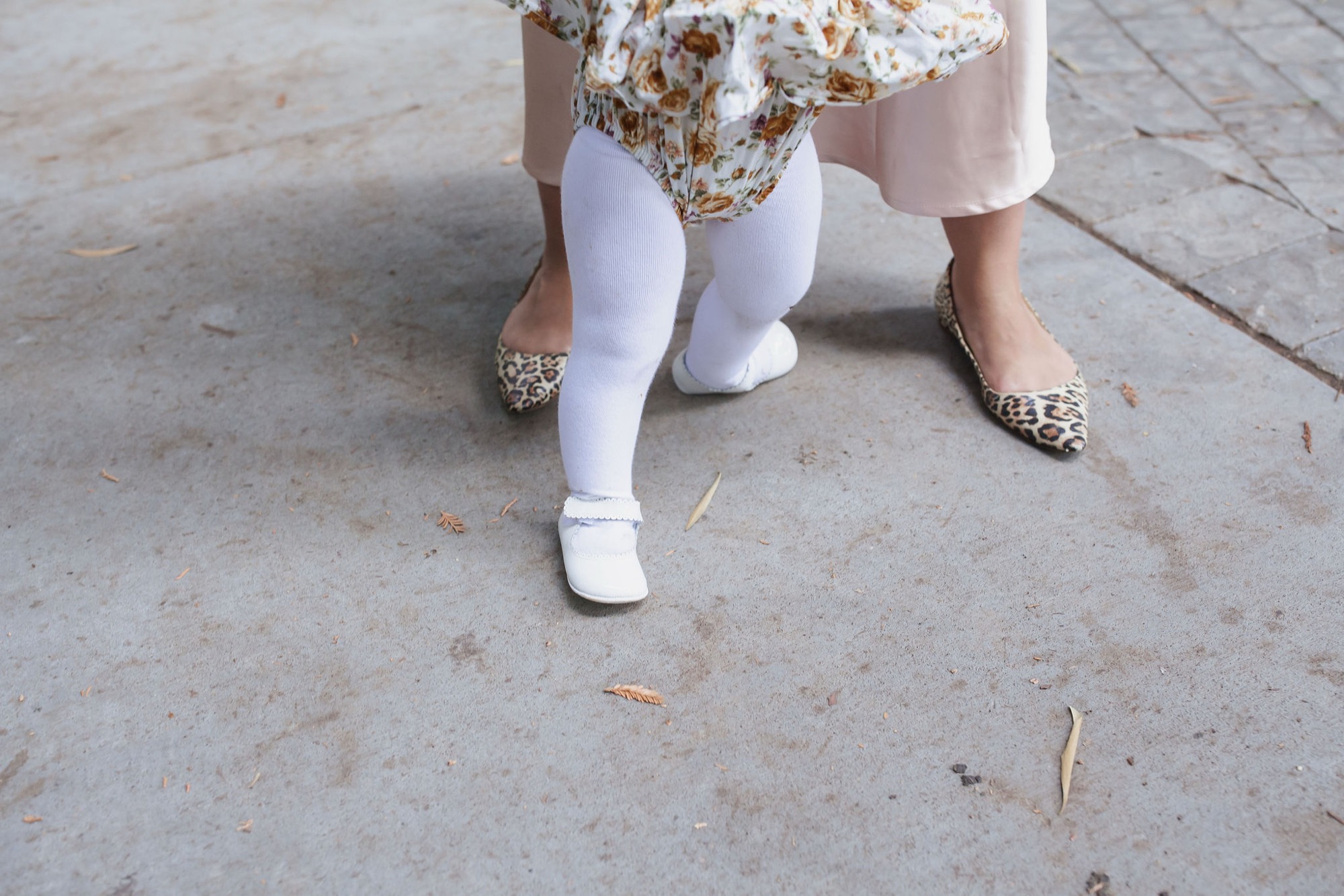 A child in white tights and sparkly shoes stands on concrete pavement, wearing a floral dress and beige cardigan