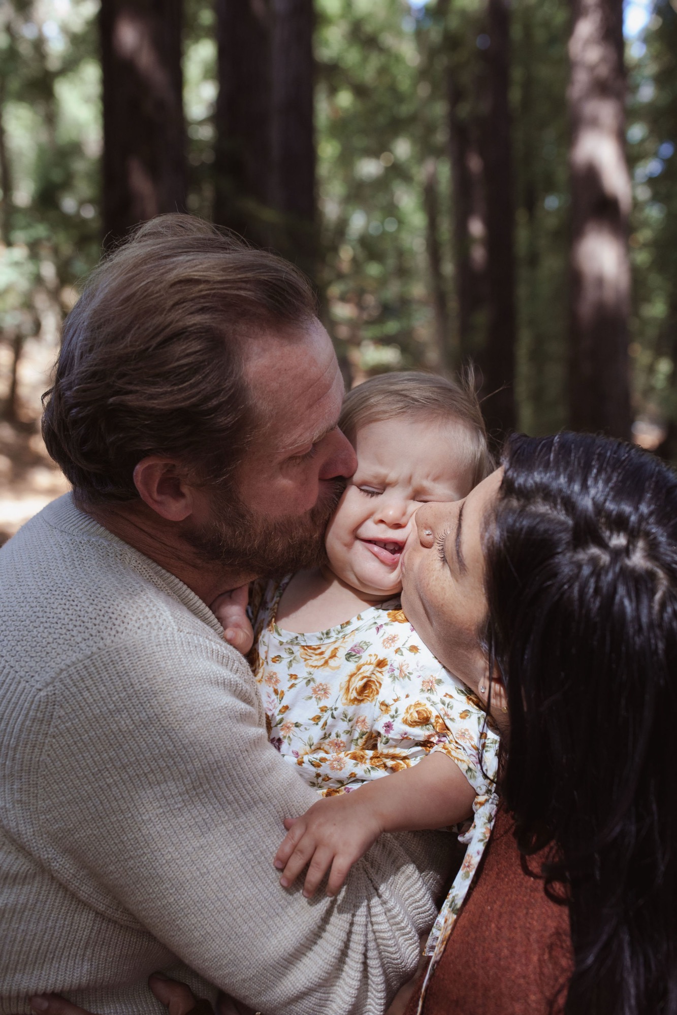 A man and woman kiss a smiling toddler between them in dappled forest light, surrounded by tall redwoods