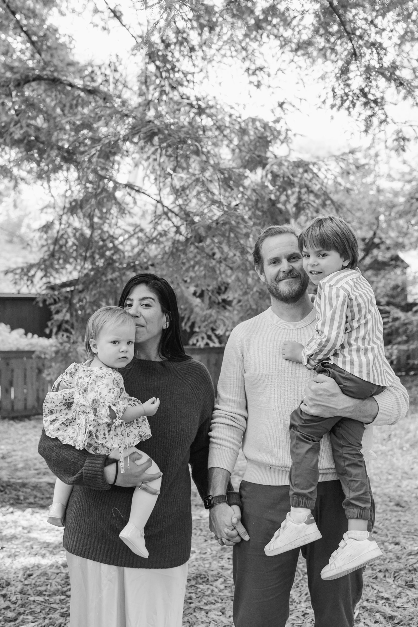 A bearded man and dark-haired woman hold their two young children beneath dappled tree shade, all gazing downward with gentle expressions