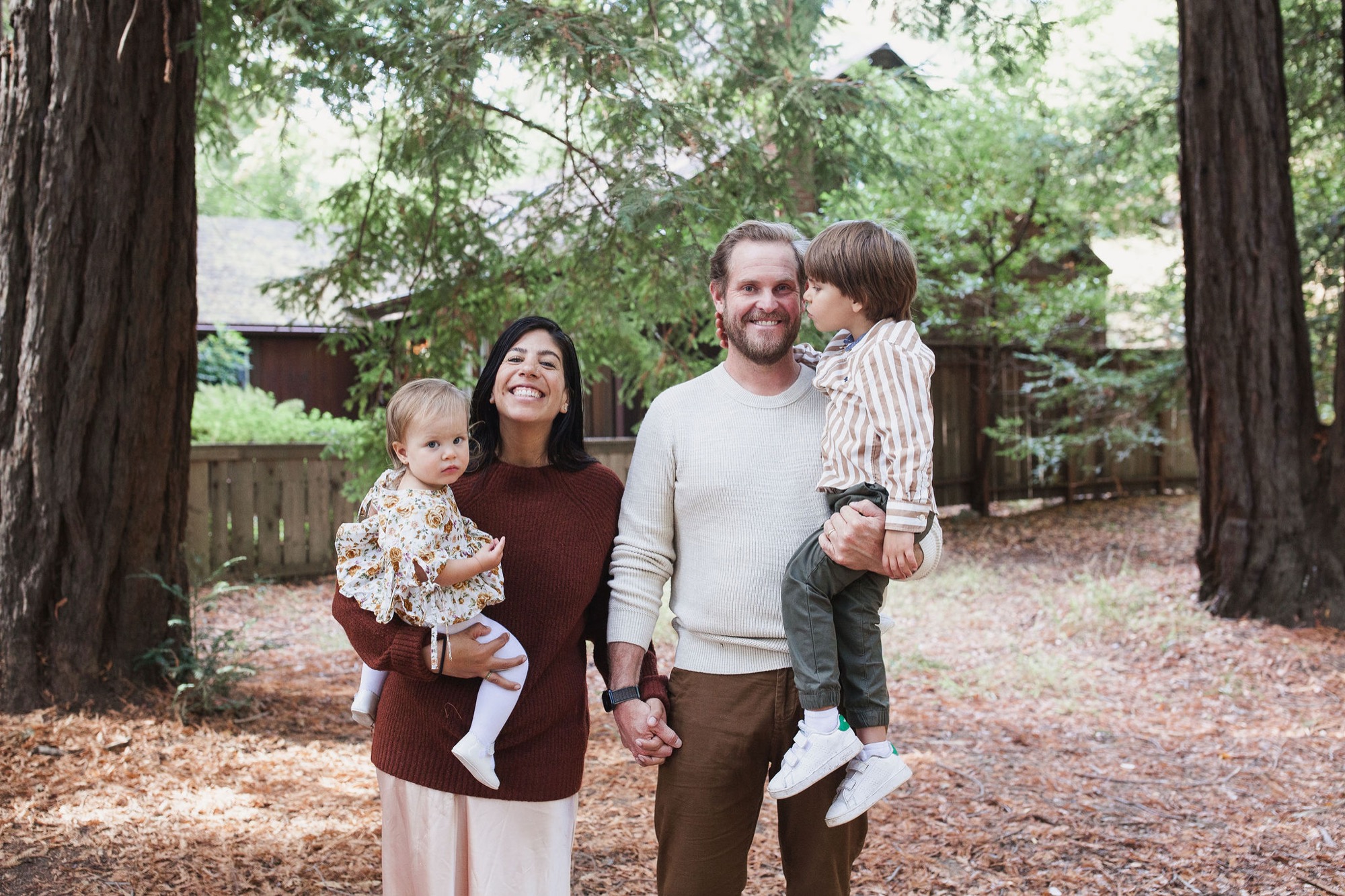 Family of four smiles warmly among towering redwoods, parents holding toddlers on a needle-covered forest floor dappled with soft afternoon light