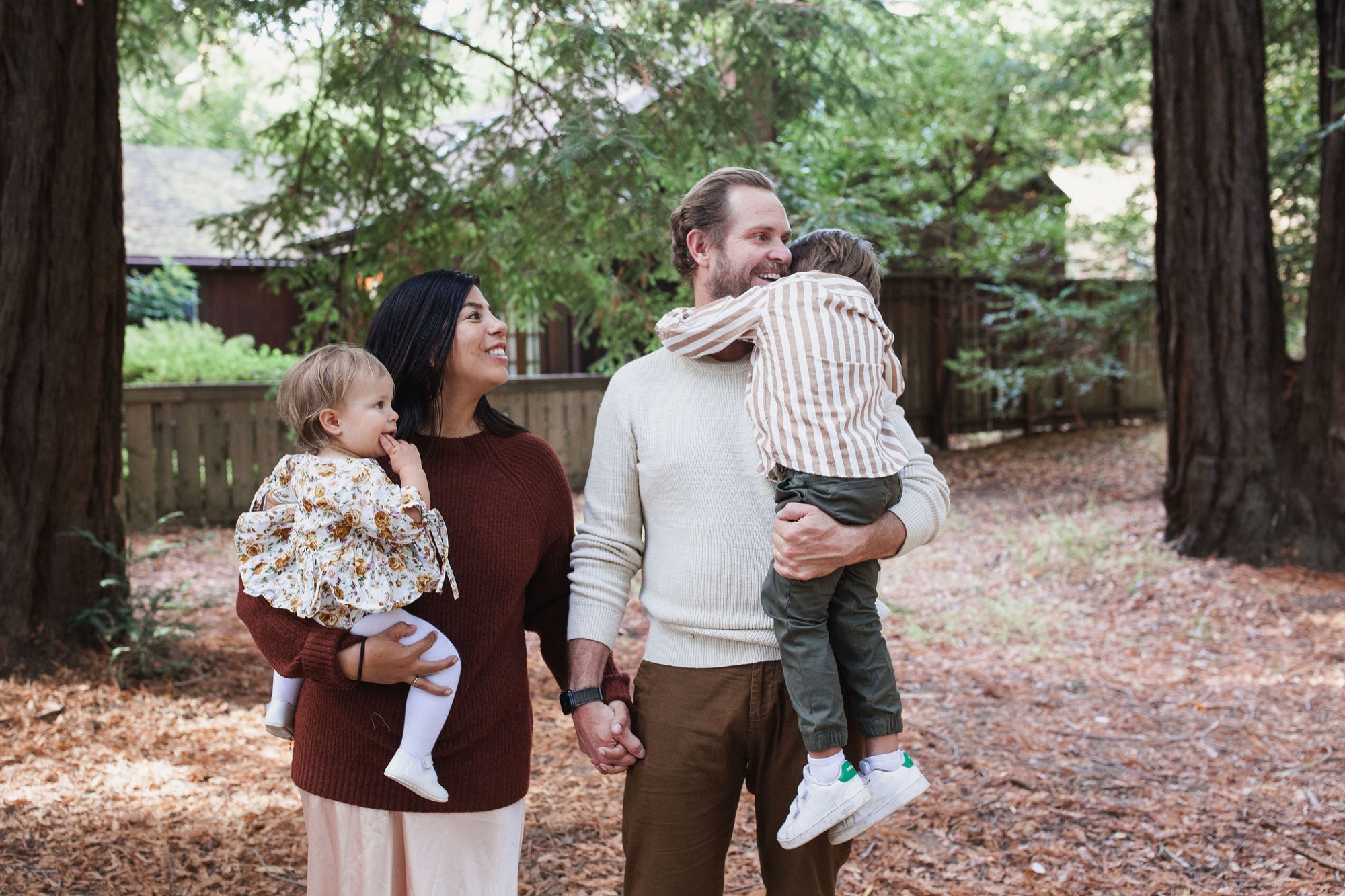 A family of four stands together on a leaf-covered forest floor beneath towering redwoods, with soft dappled sunlight filtering through the canopy