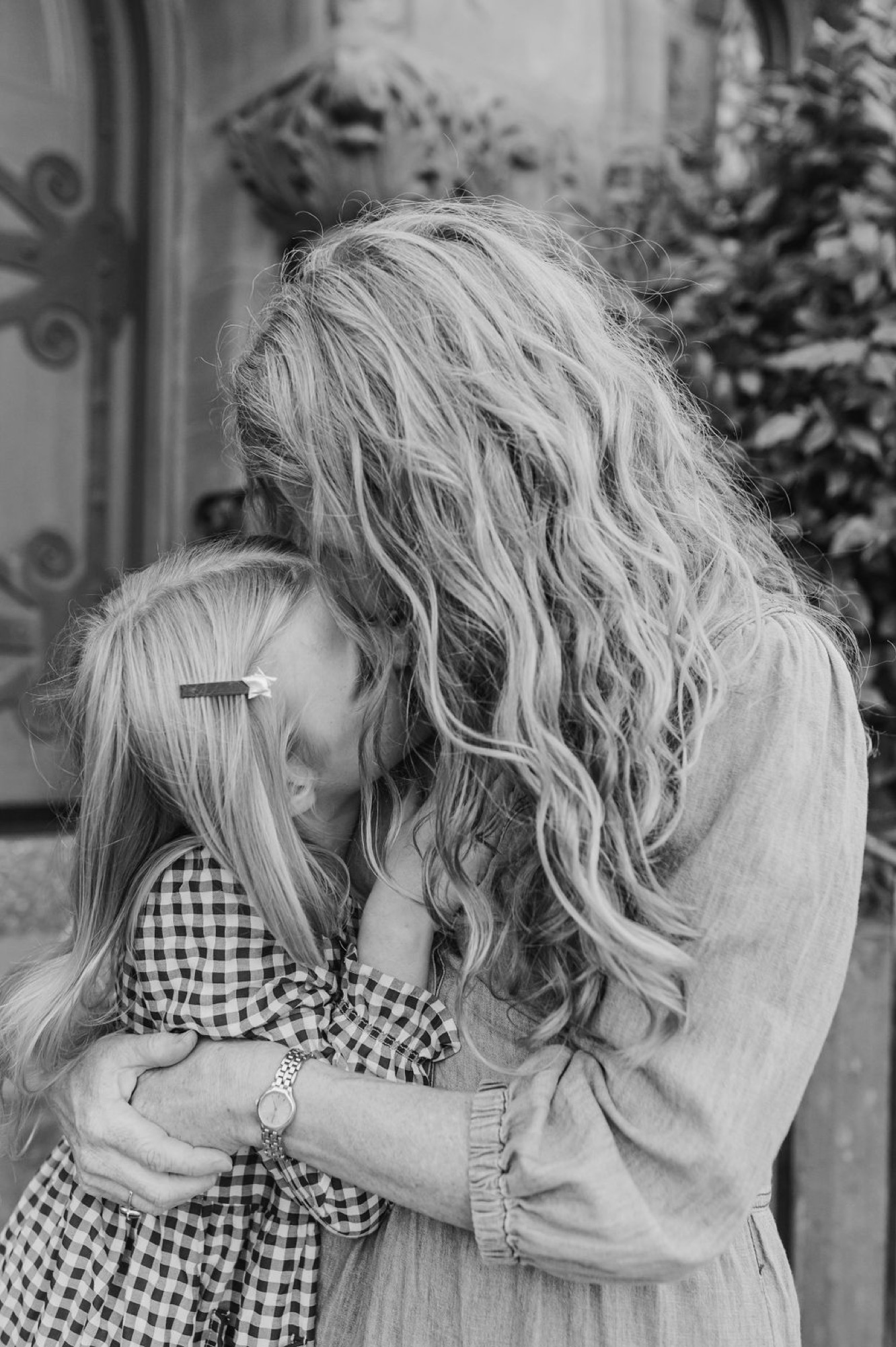 A woman in a light shirt tenderly embraces a young child in a gingham dress, their heads close together in an intimate moment against a blurred holiday backdrop — San Anselmo Family Photographer