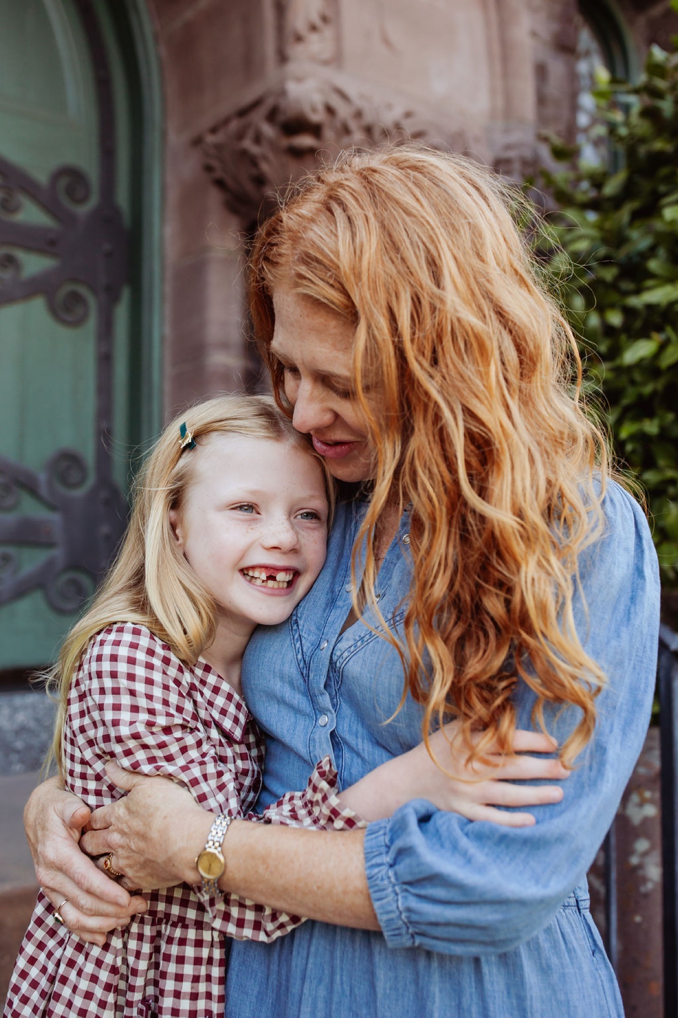 A woman with long reddish-blonde hair embraces a smiling young girl in a gingham dress against a weathered green door with ivy — San Anselmo Family Photographer