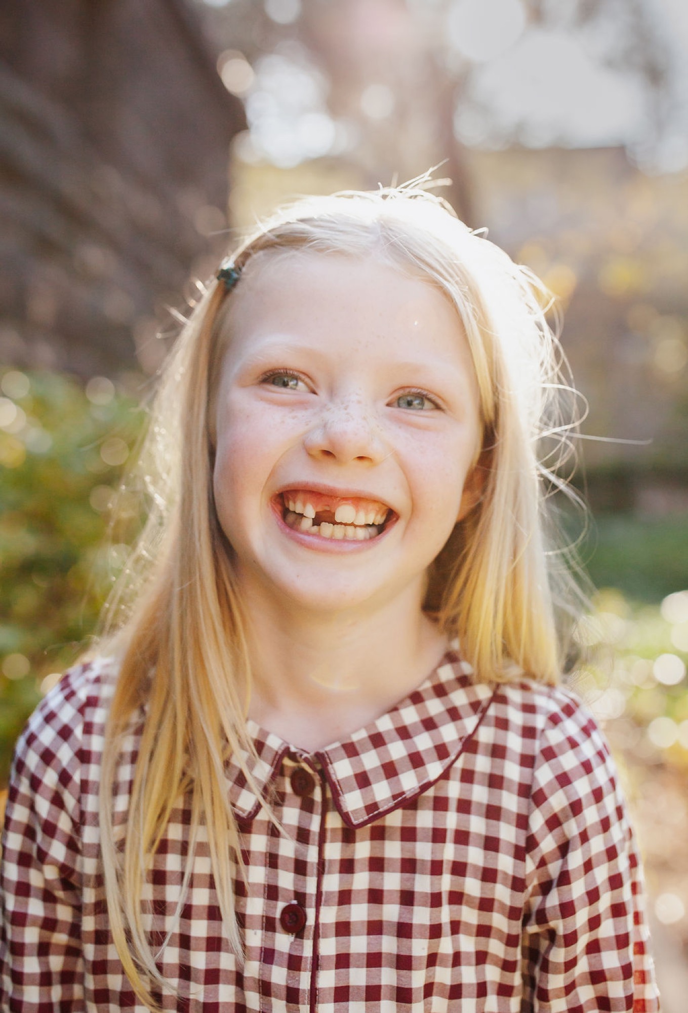 A blonde girl in a burgundy gingham shirt smiles widely at the camera with sunlit hair against a blurred outdoor background — San Anselmo Family Photographer