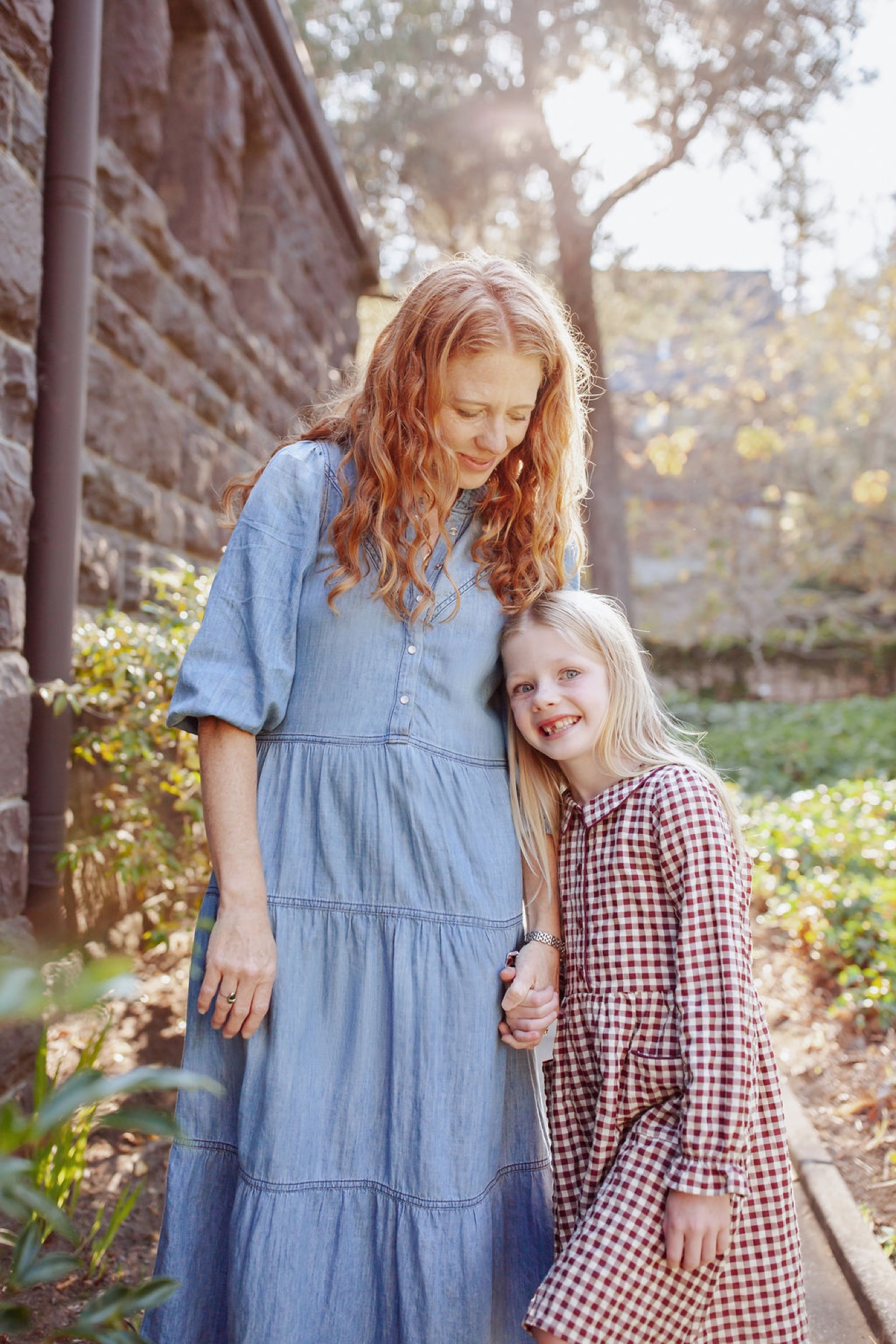 An older sister in a light blue tiered dress gazes down fondly at her younger sibling in a burgundy gingham shirt, standing close together against a brick wall with dappled sunlight filtering through overhead trees
