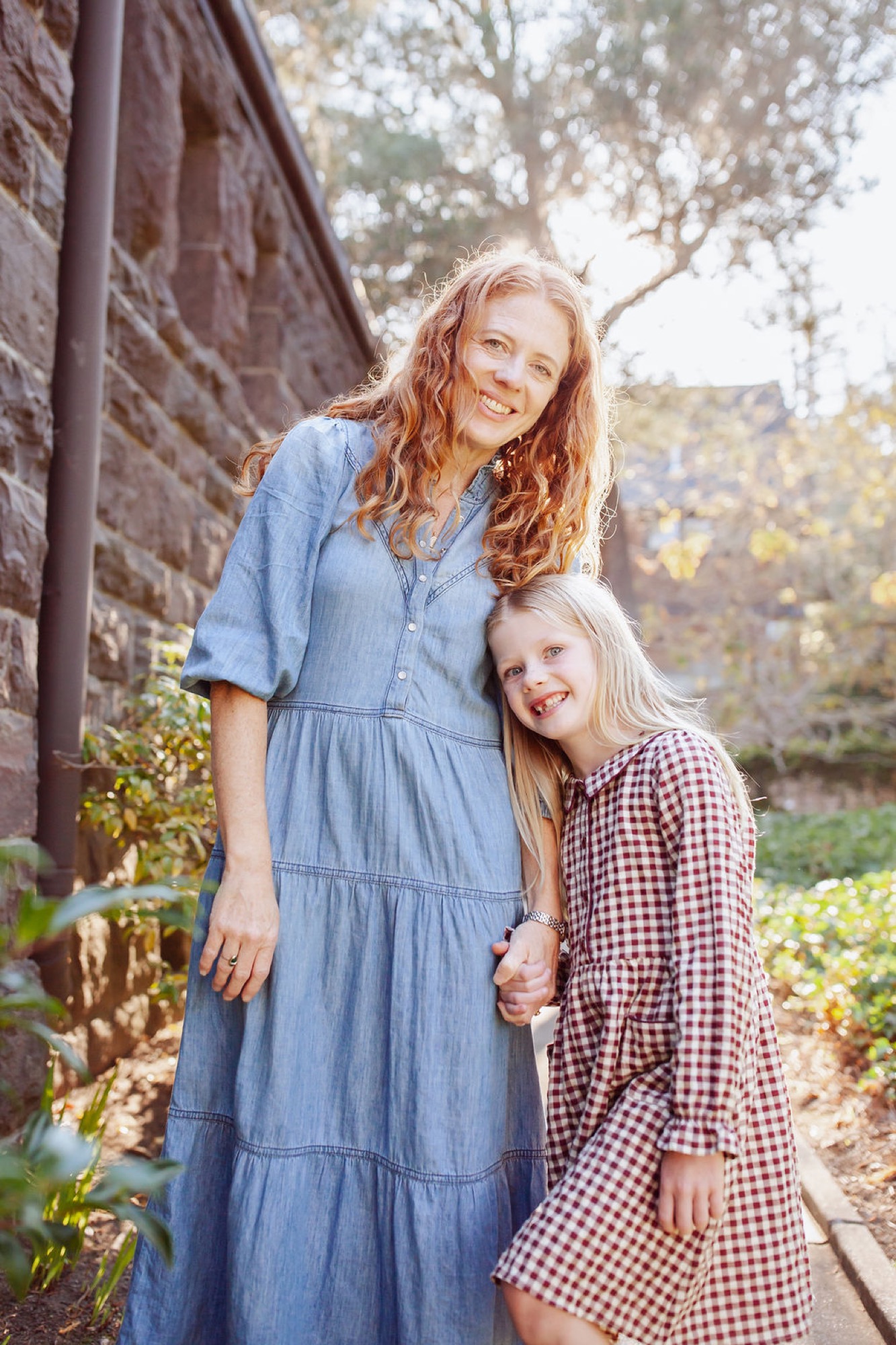 Two girls with reddish-blonde hair stand smiling beside a brick wall, dappled sunlight filtering through tree branches above them — San Anselmo Family Photographer