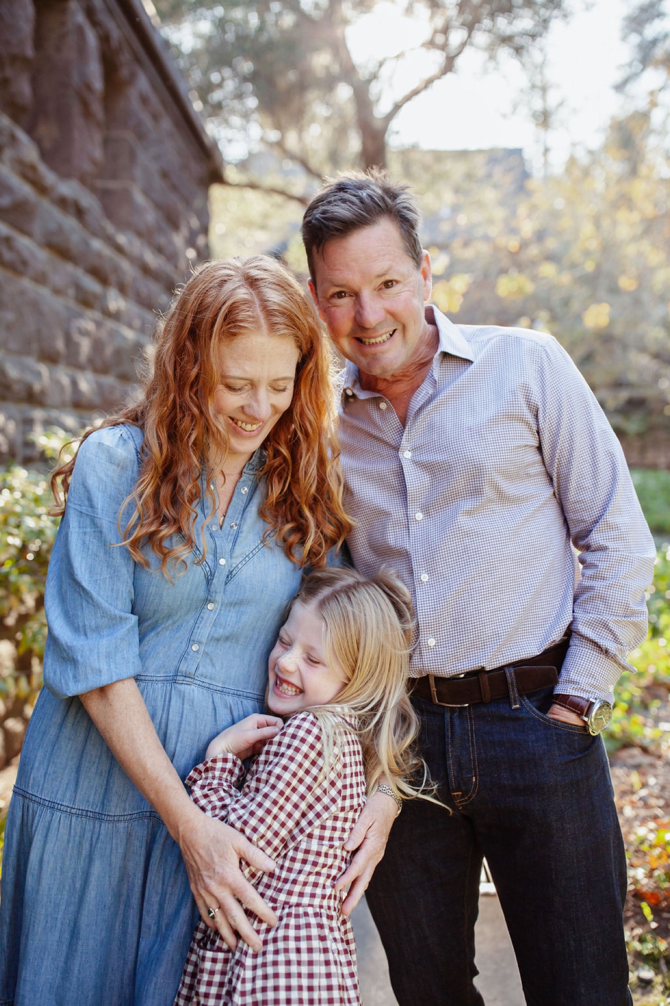 A smiling family of three stands together beneath a large tree, with the young daughter in a gingham dress nestled between her parents in soft afternoon light — San Anselmo Family Photographer