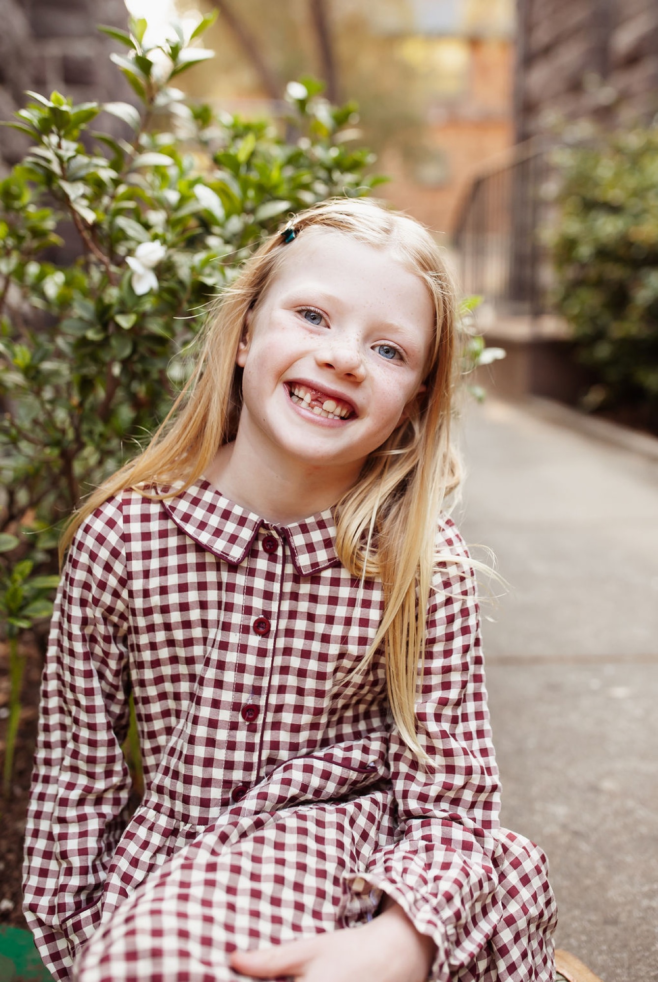 A smiling girl in a burgundy gingham shirt sits outdoors with soft afternoon light illuminating her genuine expression and long blonde hair — San Anselmo Family Photographer