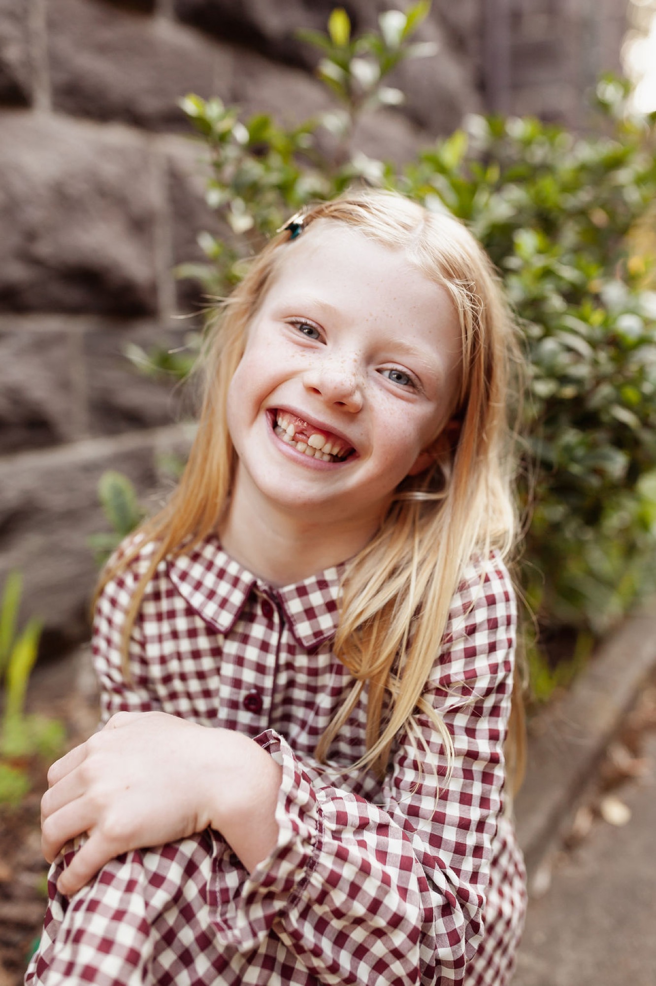 A young girl in a burgundy gingham shirt laughs broadly at the camera with genuine delight, standing before a textured stone wall and green foliage — San Anselmo Family Photographer