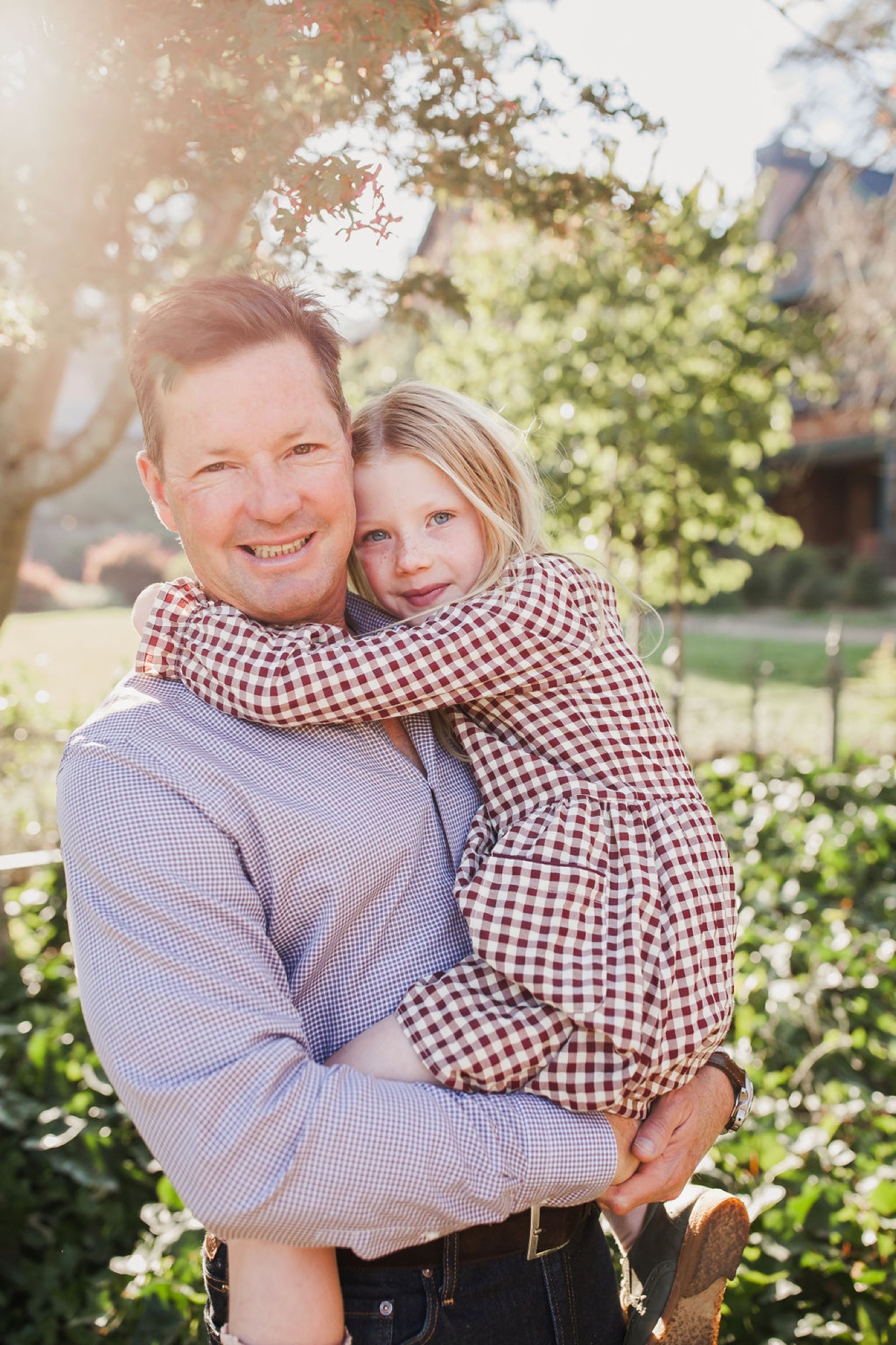 A man in a blue gingham shirt holds his young daughter wearing a brown checkered dress, both smiling warmly in dappled sunlight beneath flowering trees — San Anselmo Family Photographer