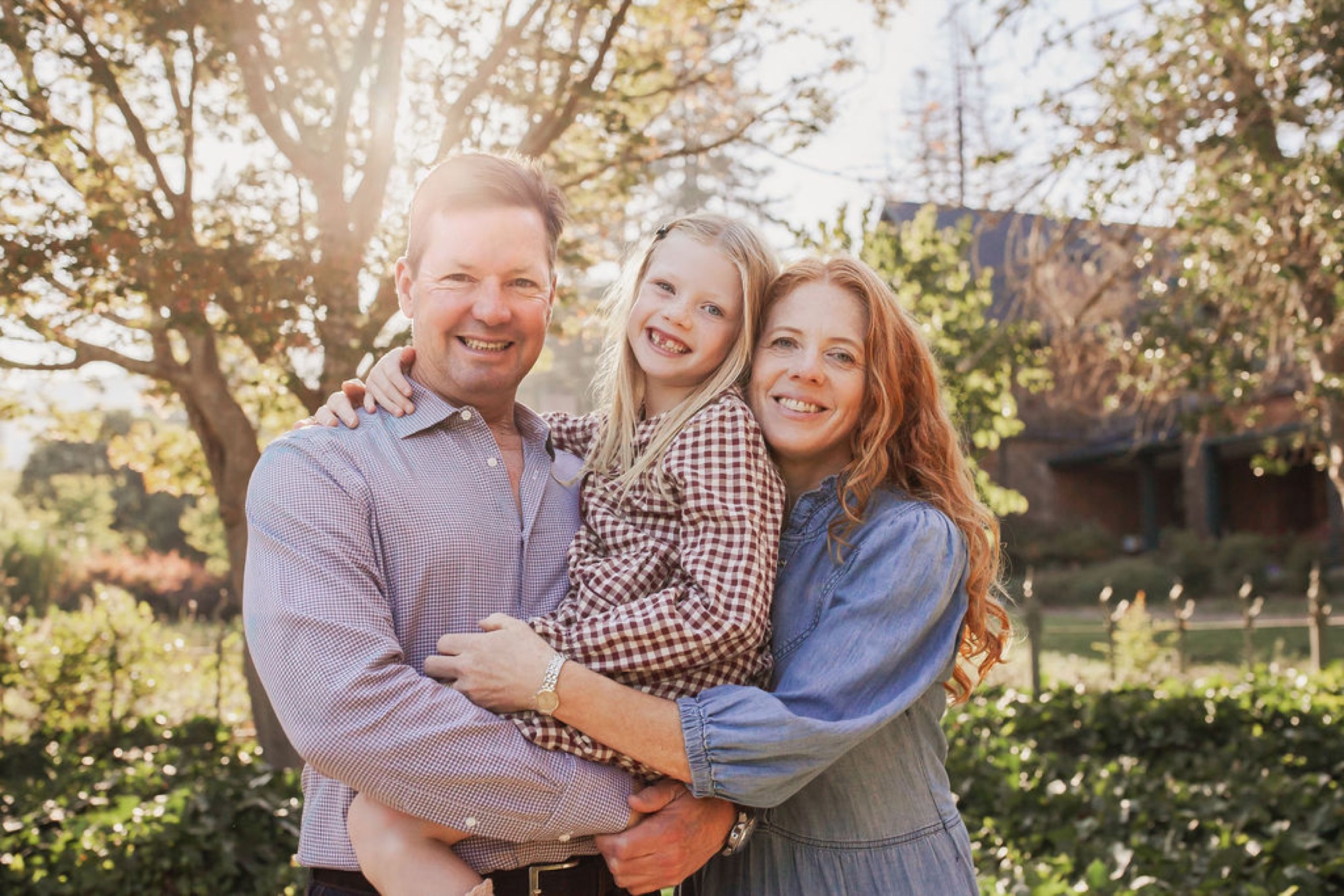 A smiling family of three stands together beneath golden-lit trees, with the father holding his young daughter while the mother embraces them both from the side — San Anselmo Family Photographer