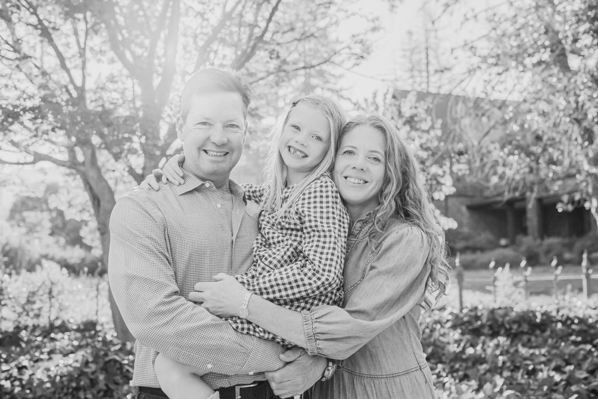 A man and woman embrace their young daughter between them in a sun-dappled garden, all three beaming with genuine joy — San Anselmo Family Photographer