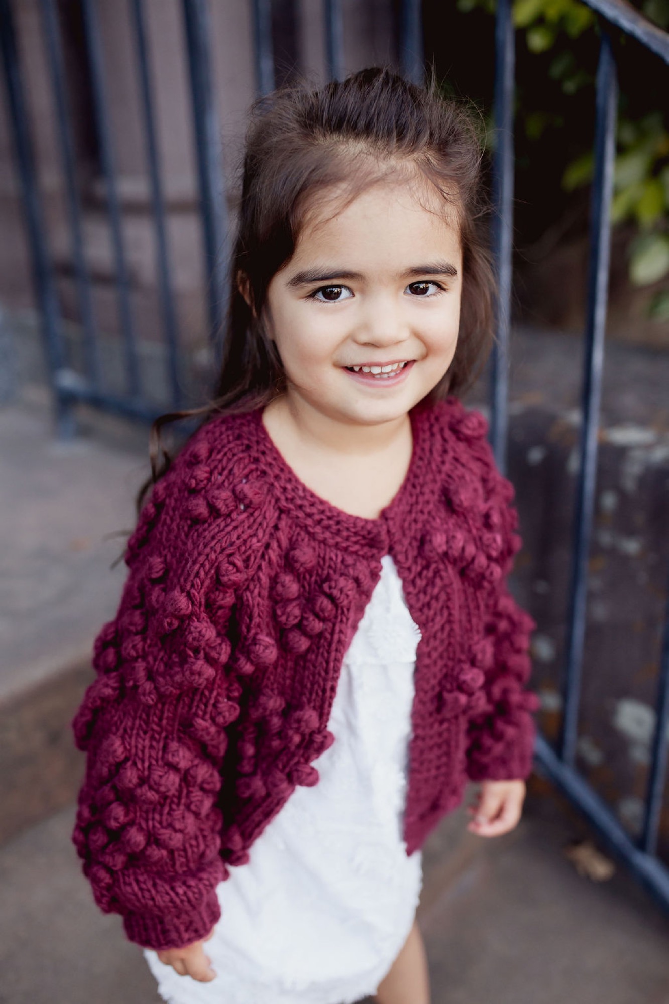 Young girl in burgundy knit cardigan smiles brightly at camera on a downtown street with blurred storefront backdrop