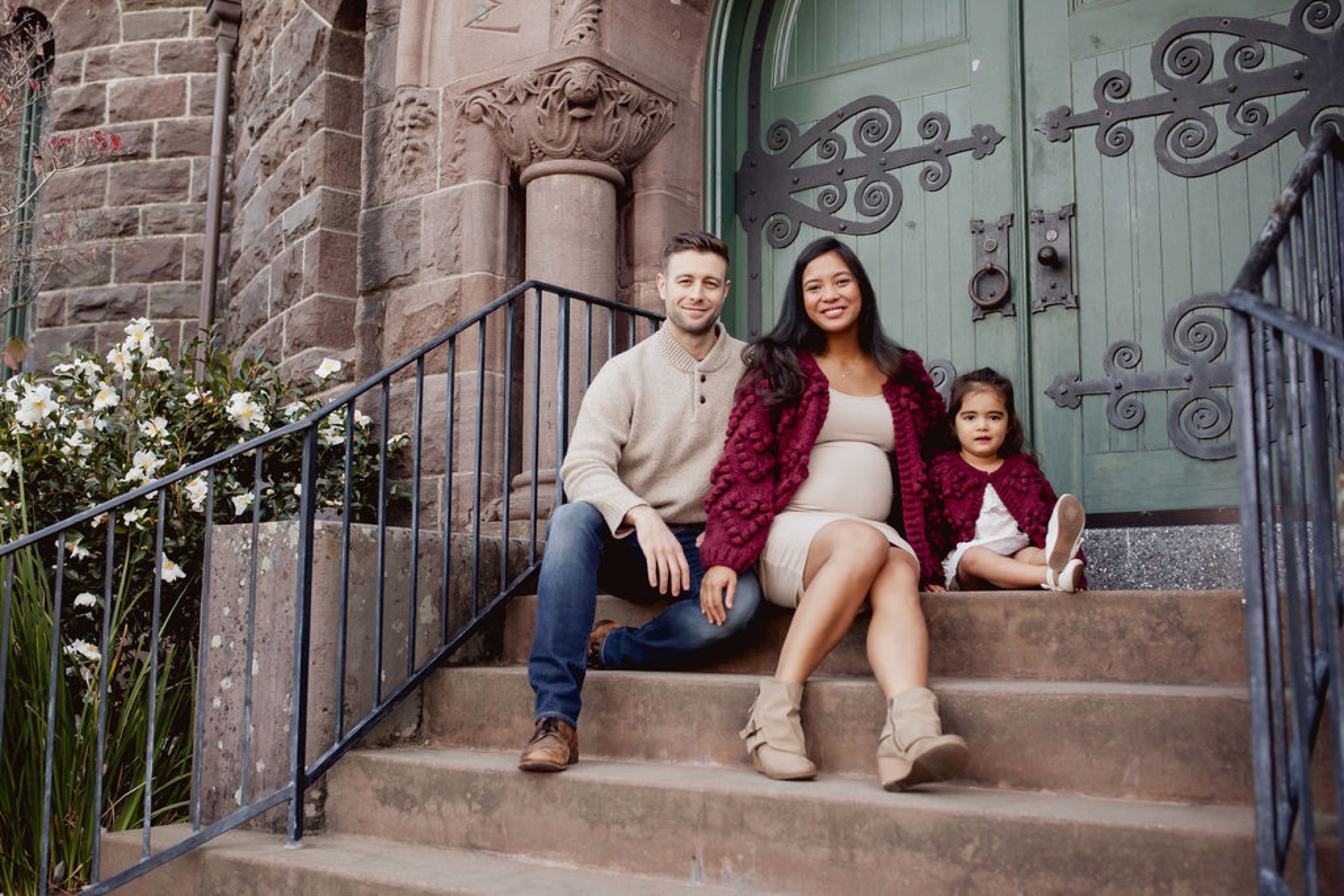 A family of three sits together on brick steps beside an ornate green door, with the mother in the center smiling warmly while the father and young daughter lean in close — San Anselmo Family Photographer