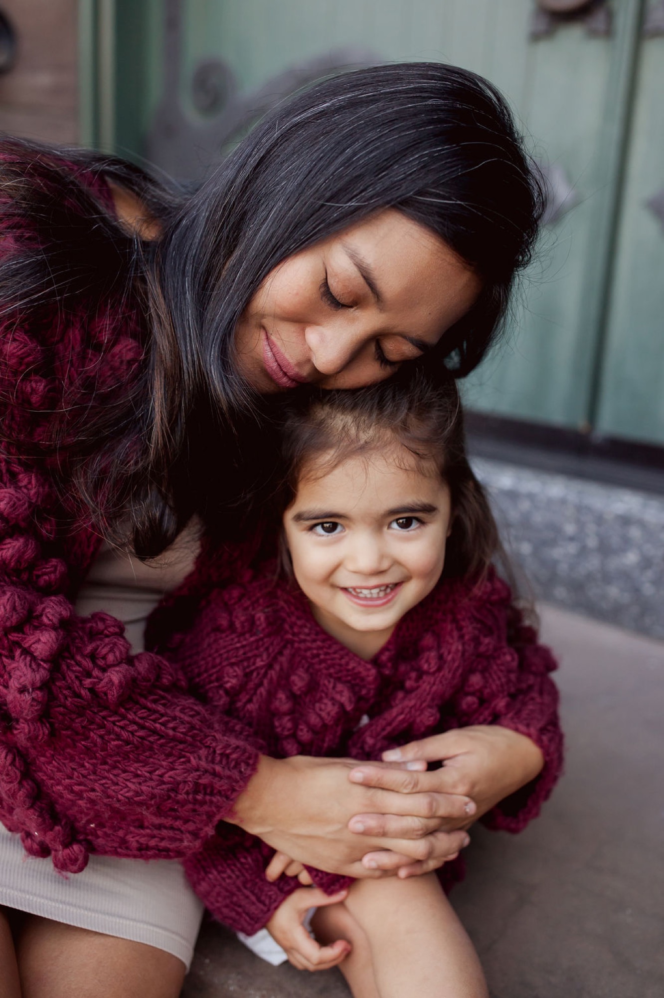 A mother in burgundy embraces her young daughter from behind, both wearing matching maroon sweaters, with genuine smiles and eyes closed in tender affection — San Anselmo Family Photographer