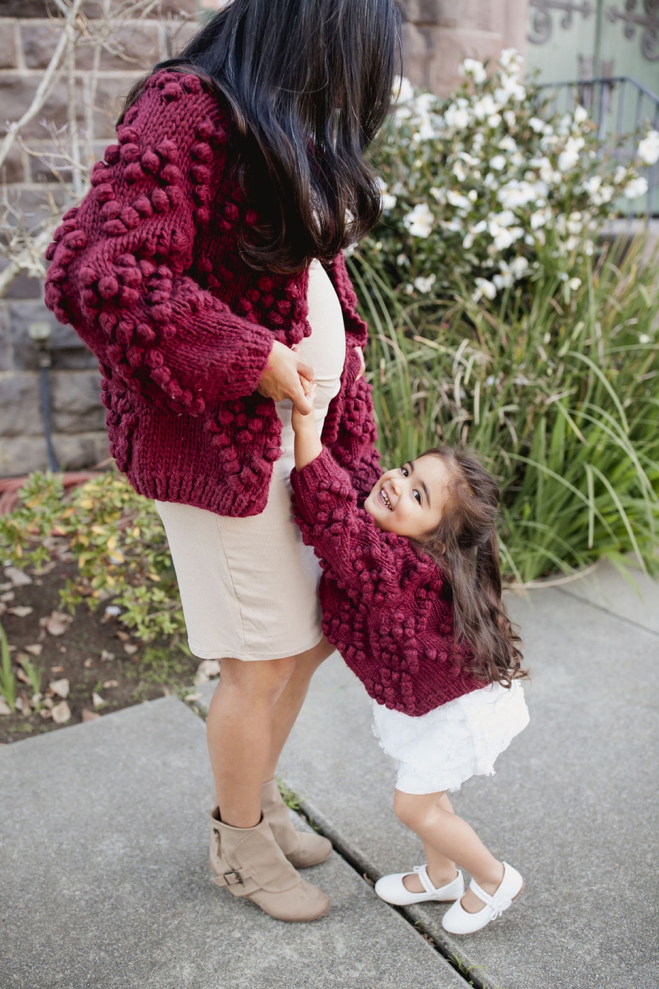 A mother and young daughter stand together on a sidewalk wearing matching burgundy cardigans, the child gazing up with a gentle smile