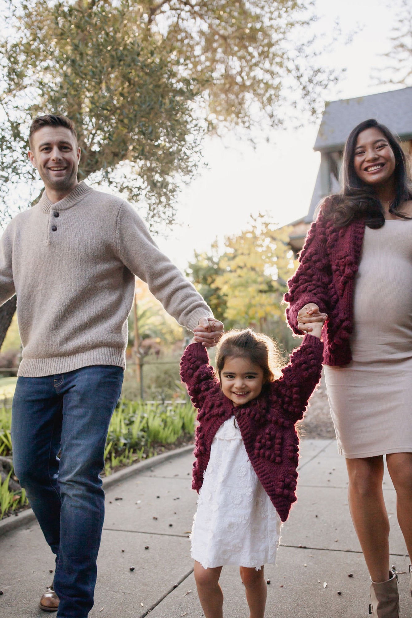 A family of three walks hand-in-hand down a concrete path lined with autumn trees, with the child swinging between her parents' arms — San Anselmo Family Photographer