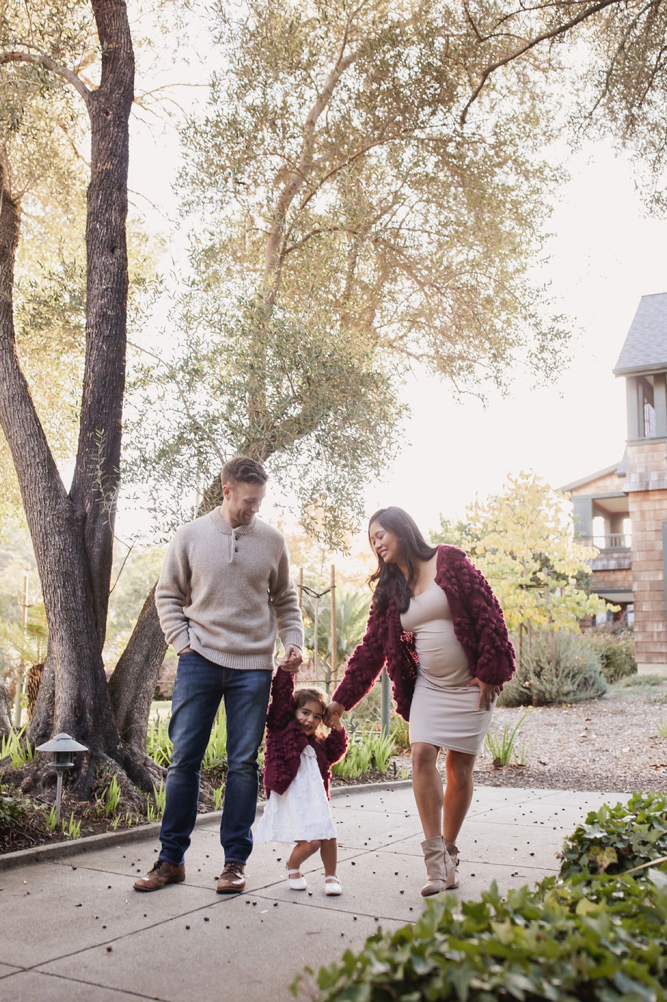Parents walk hand-in-hand with their young daughter along a shaded pathway beneath a large tree, autumn light filtering through sparse foliage