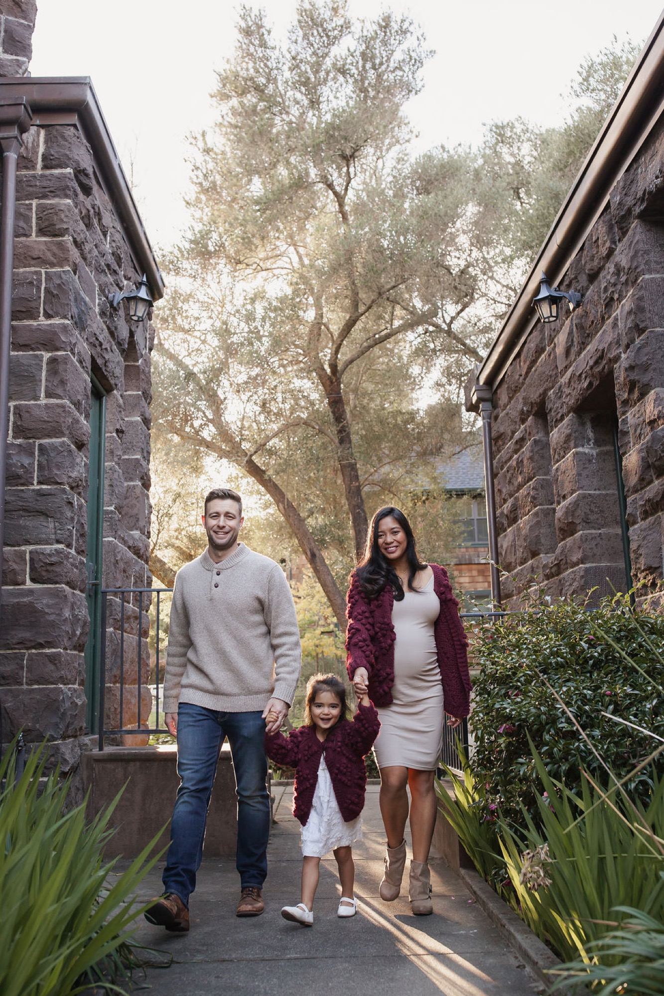 A family of three walks together down a narrow brick pathway between stone buildings, with dappled sunlight filtering through an overhead tree