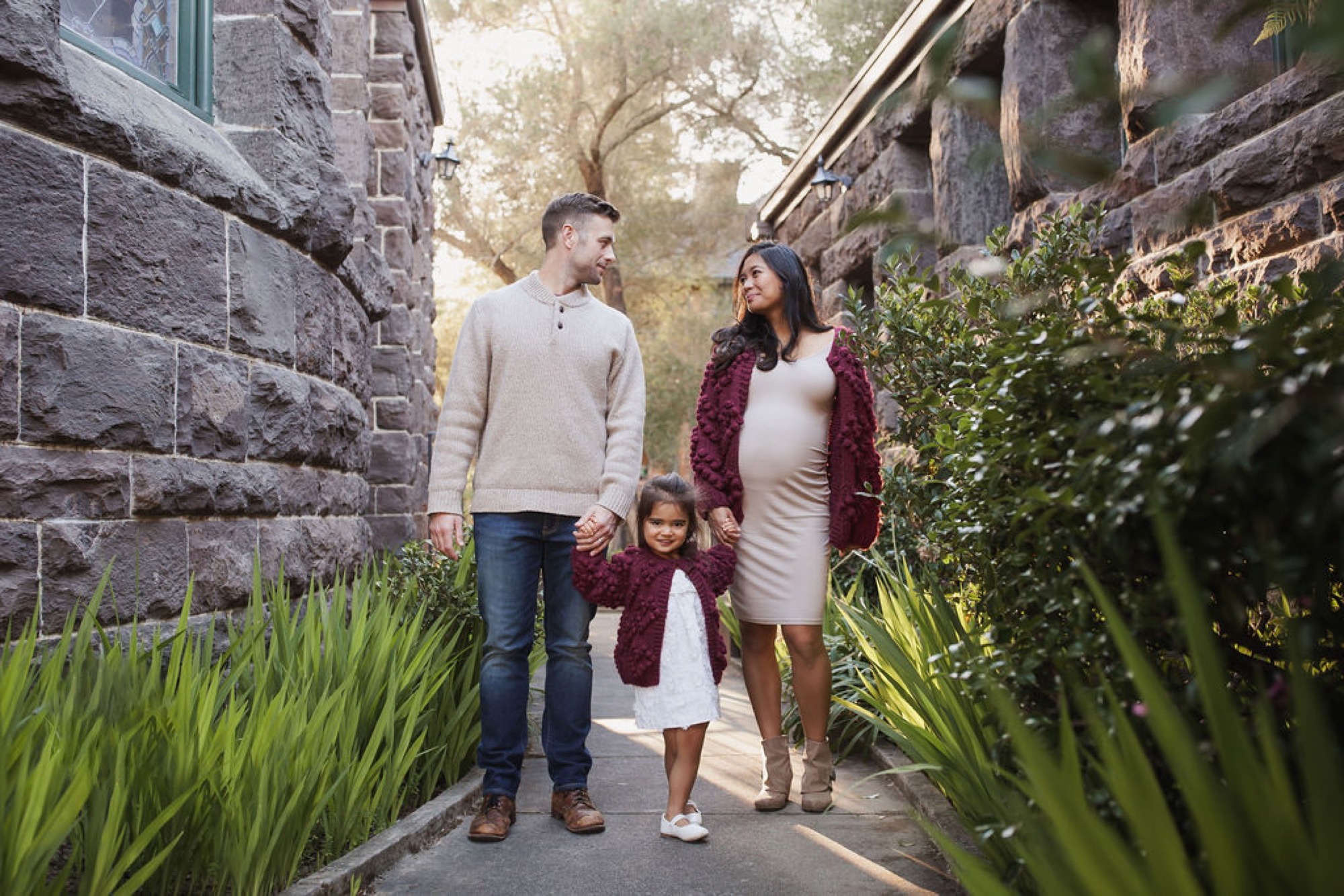 A family of three walks hand-in-hand down a narrow stone pathway lined with lush green plants, dressed in complementary burgundy and cream tones — San Anselmo Family Photographer