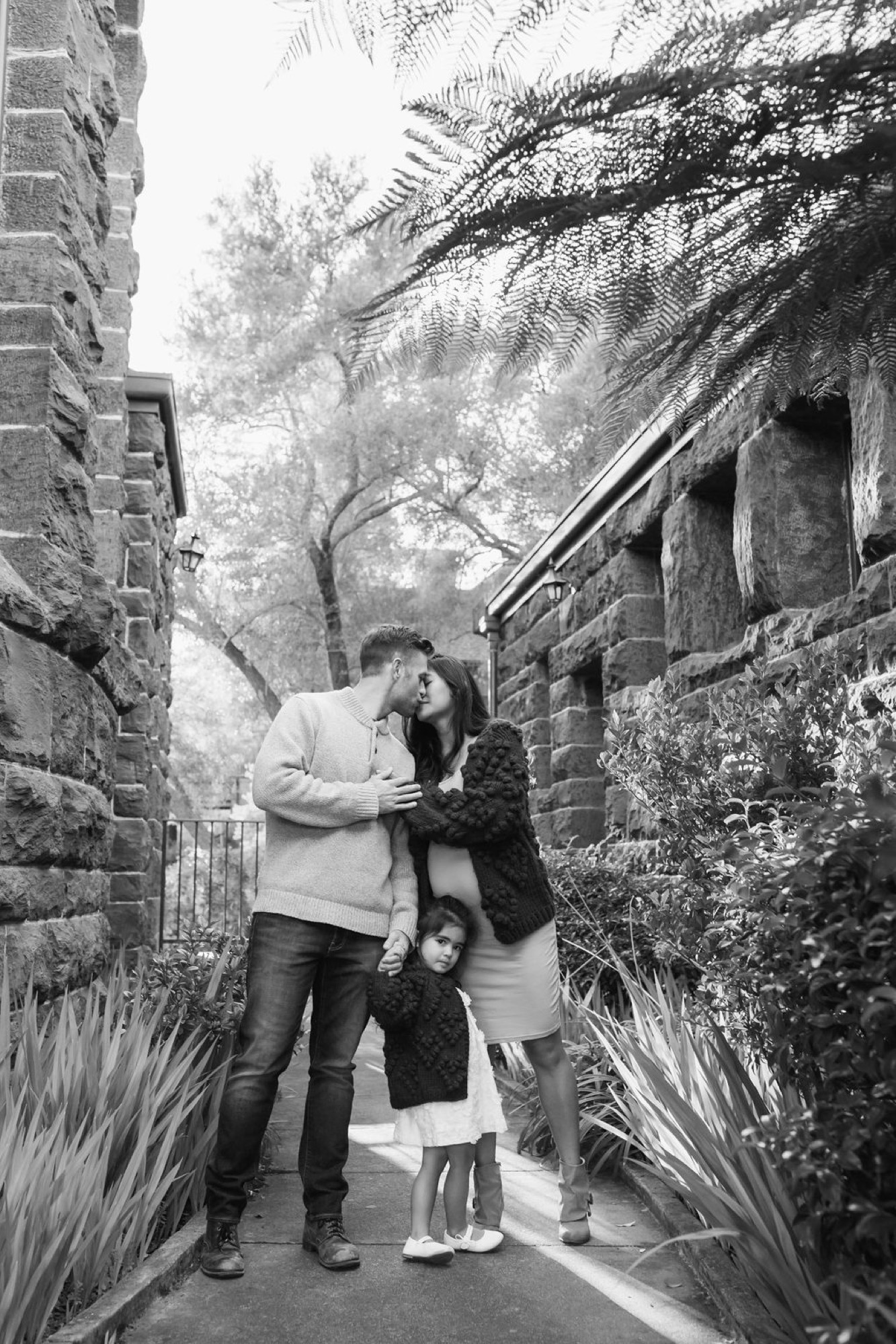 A father bends down to embrace his two young children on a narrow stone pathway lined with ivy-covered walls
