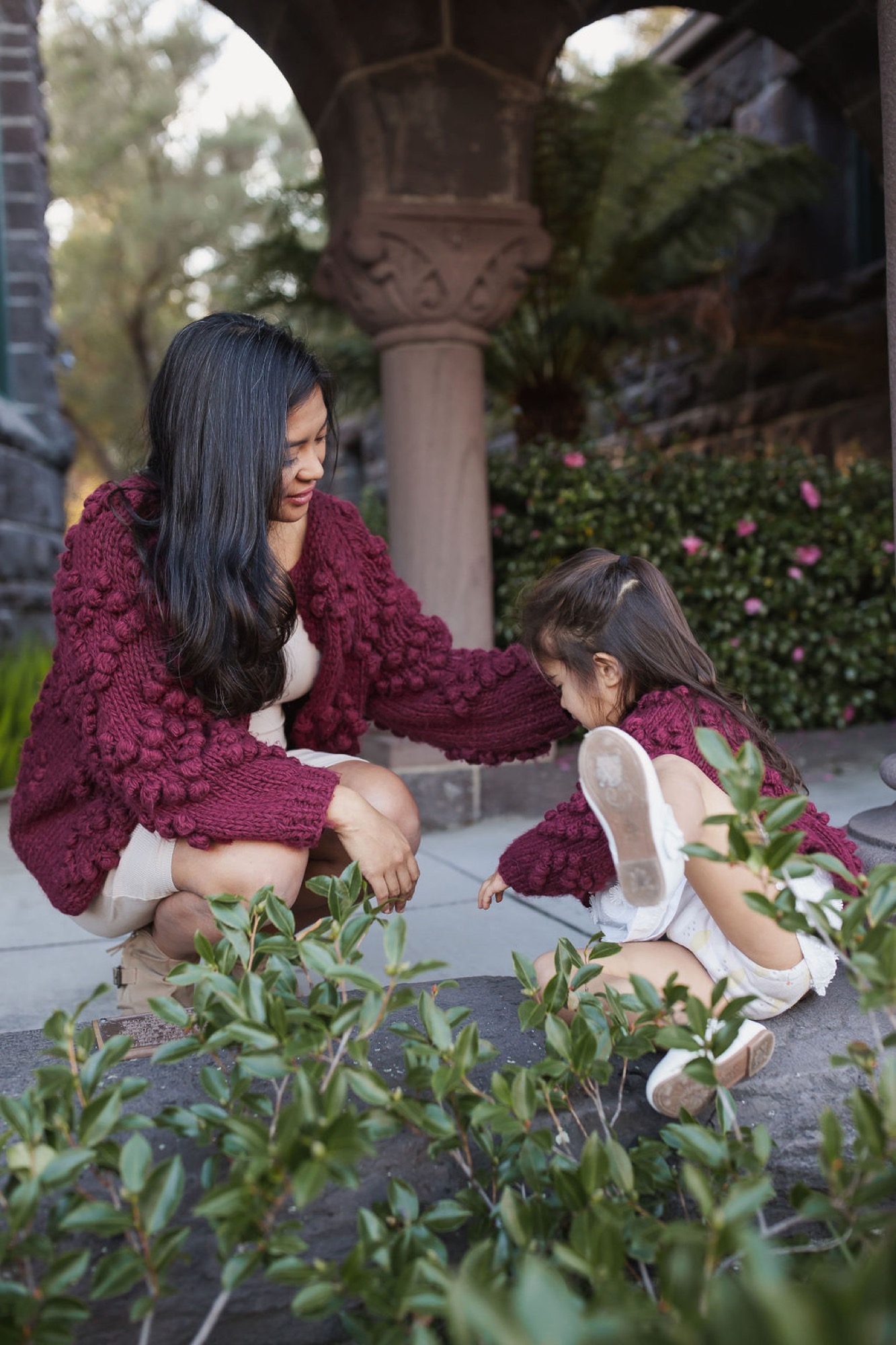 A woman in a burgundy cardigan crouches beside a young child in matching maroon, both examining green plants together under a leafy archway