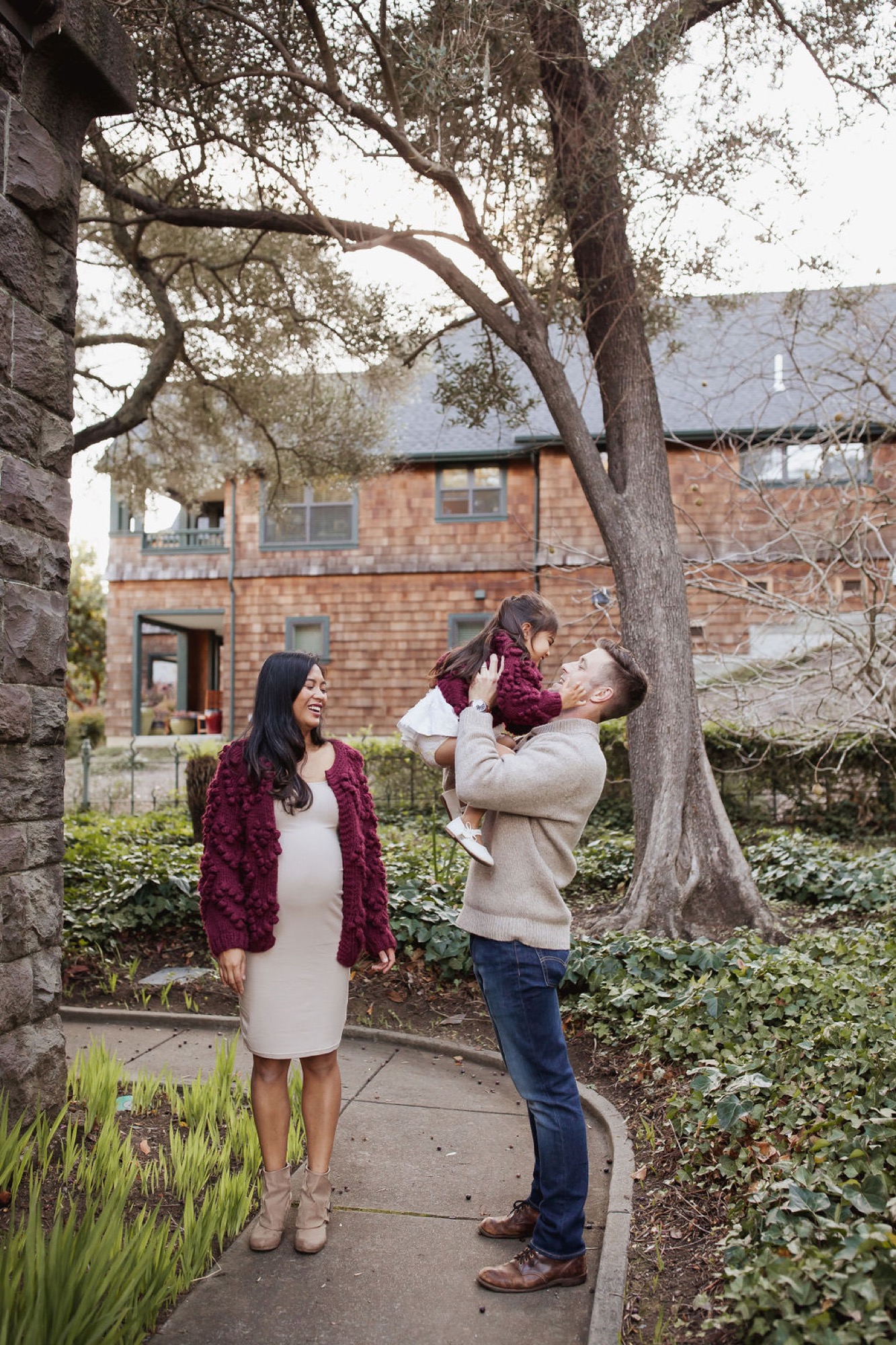 Father tosses his toddler playfully while mother watches beside him under a sprawling oak tree in a brick courtyard