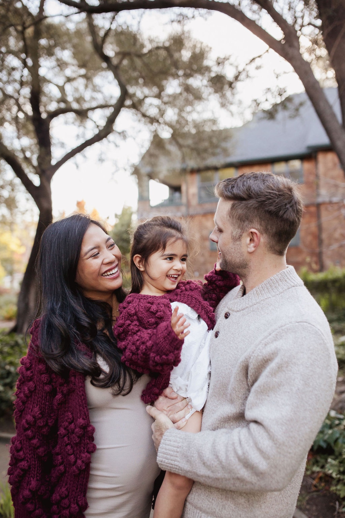 Parents and young daughter smile warmly beneath a bare oak tree, the mother glowing in a burgundy wrap and cream dress while father holds their child