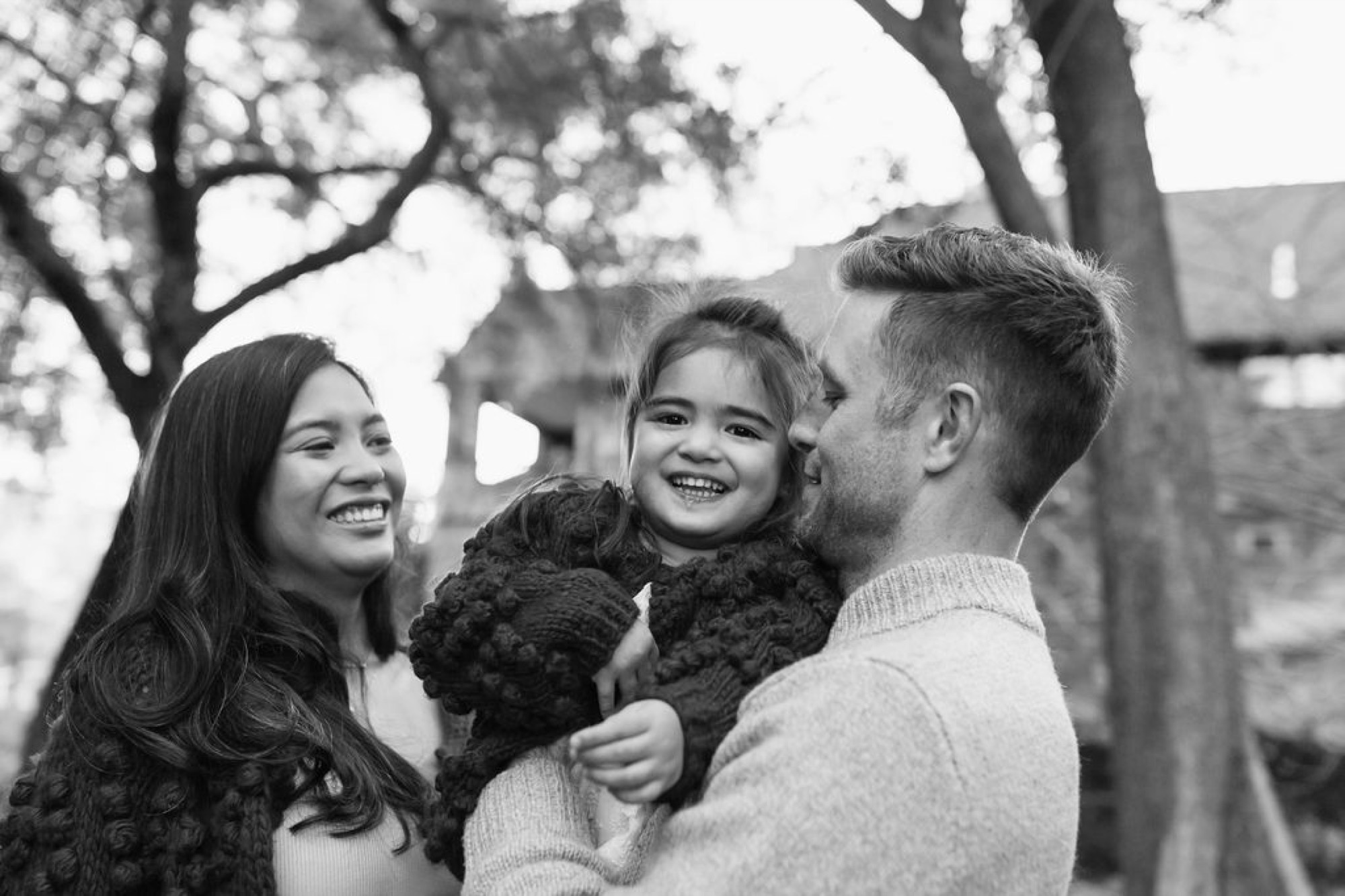 A young family laughs together outdoors, the father holding their smiling daughter while the mother gazes warmly at them both beneath bare oak trees — San Anselmo Family Photographer