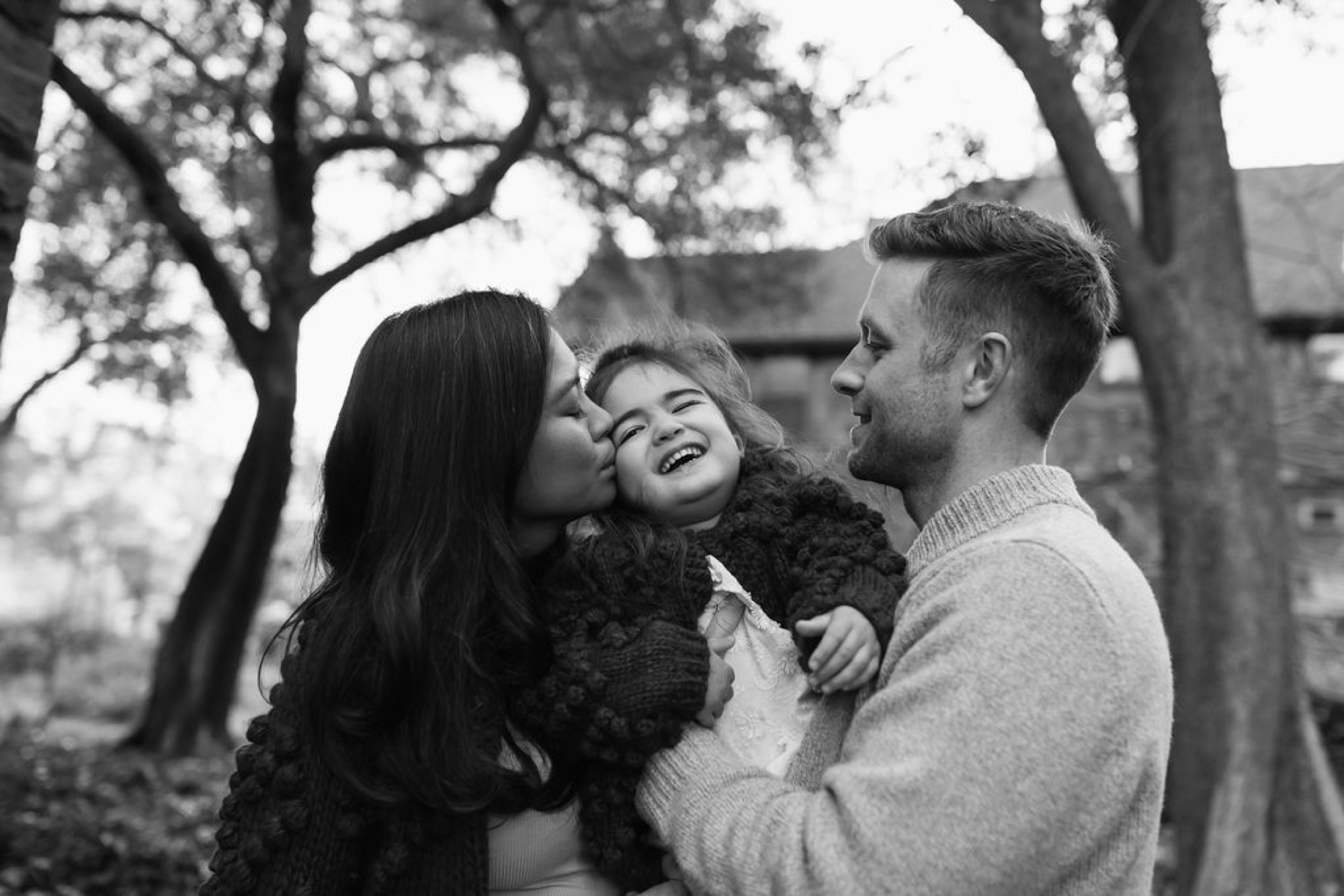 A young child laughs joyfully while held between two parents beneath bare oak trees in soft winter light