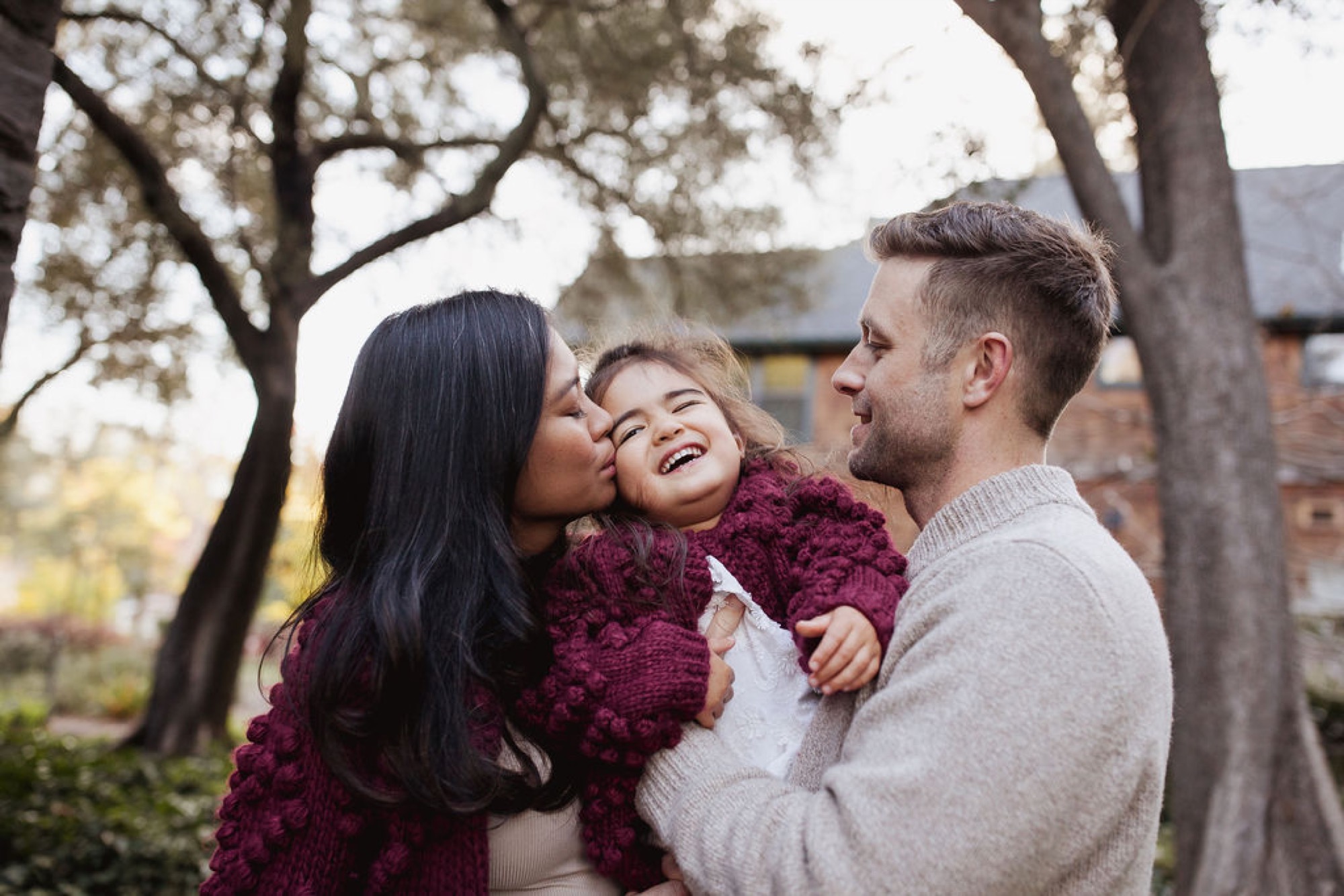 A mother kisses her young daughter's cheek while the father smiles beside them under bare oak trees in soft afternoon light