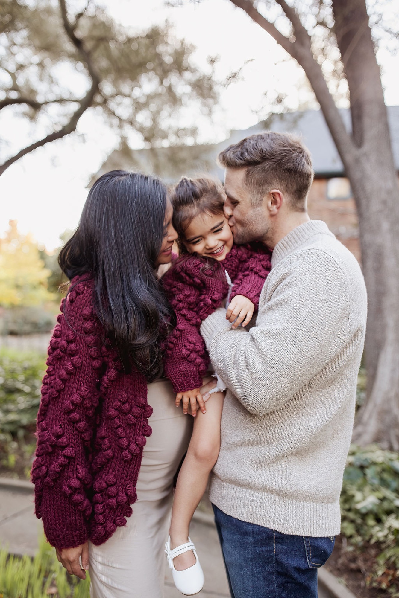 Parents kiss their toddler's cheeks while holding her between them under sprawling oak trees, soft natural light filtering through branches