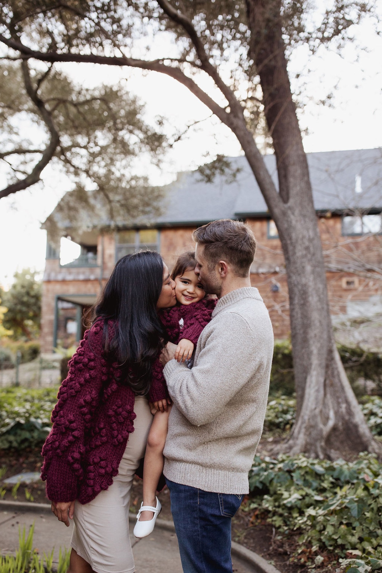 Parents hold their young daughter beneath a sprawling oak tree, her burgundy dress matching her mother's as soft afternoon light filters through the branches — San Anselmo Family Photographer