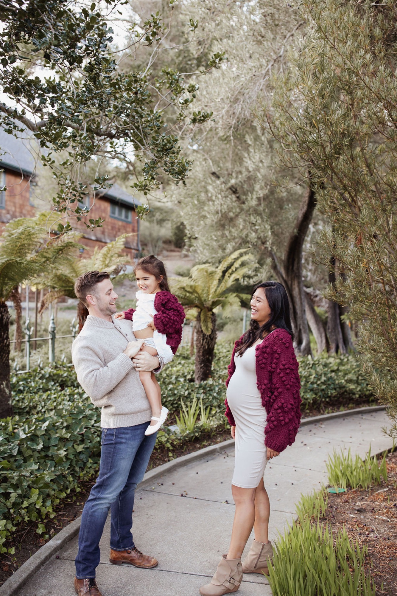 A man holding a toddler in a burgundy dress converses with a pregnant woman on a tree-lined downtown pathway