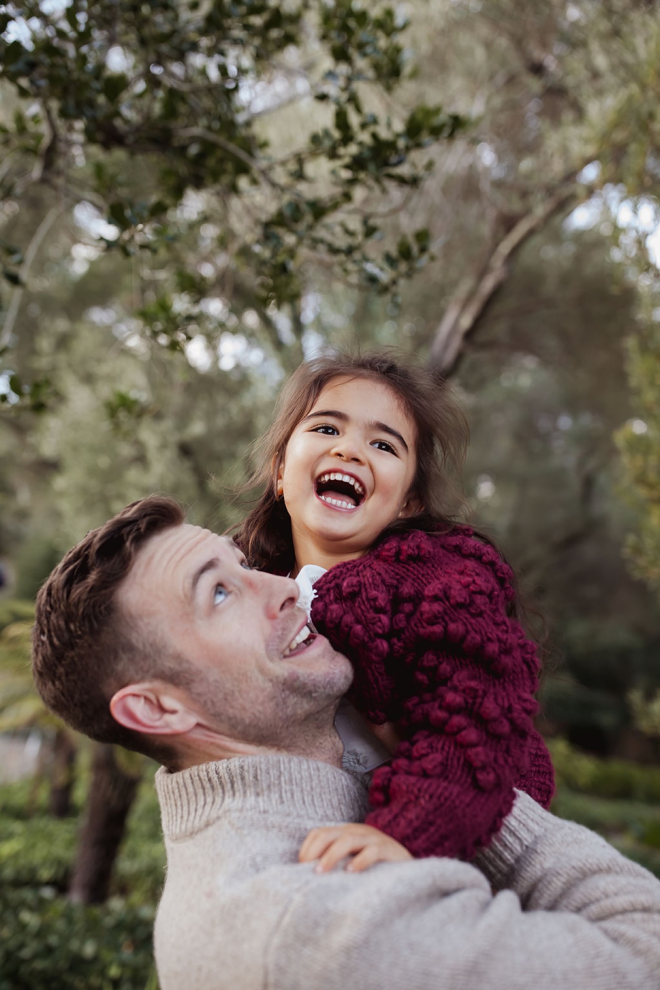 A man lifts a laughing child in a burgundy sweater beneath eucalyptus trees, both gazing upward with genuine joy — San Anselmo Family Photographer