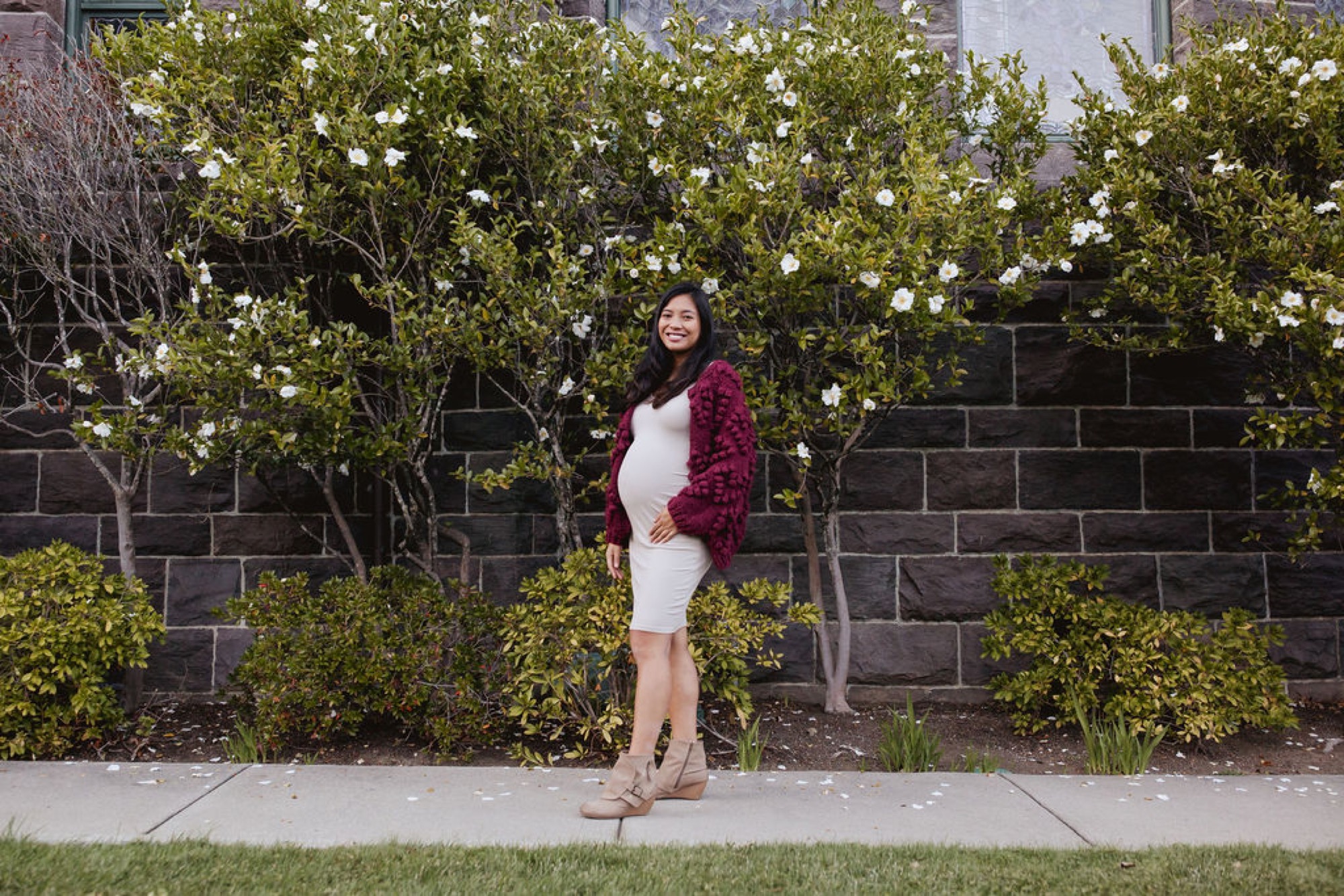 Pregnant woman in burgundy cardigan and white dress stands confidently against a dark brick wall with blooming white flowers