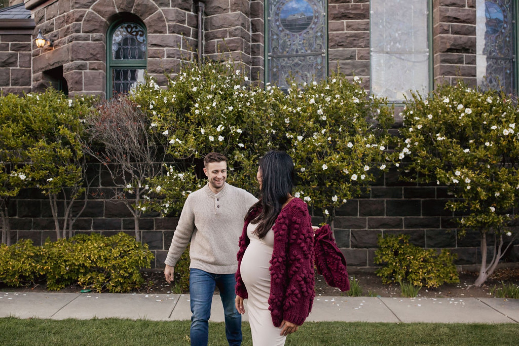 Expecting parents walk together past blooming white flowers in front of a historic stone building, the man in a cream sweater and the woman cradling her belly
