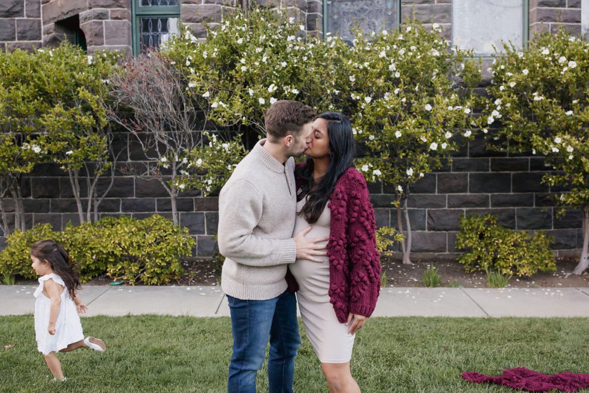 A couple shares an intimate kiss while their young daughter runs playfully across the manicured lawn in front of a brick building
