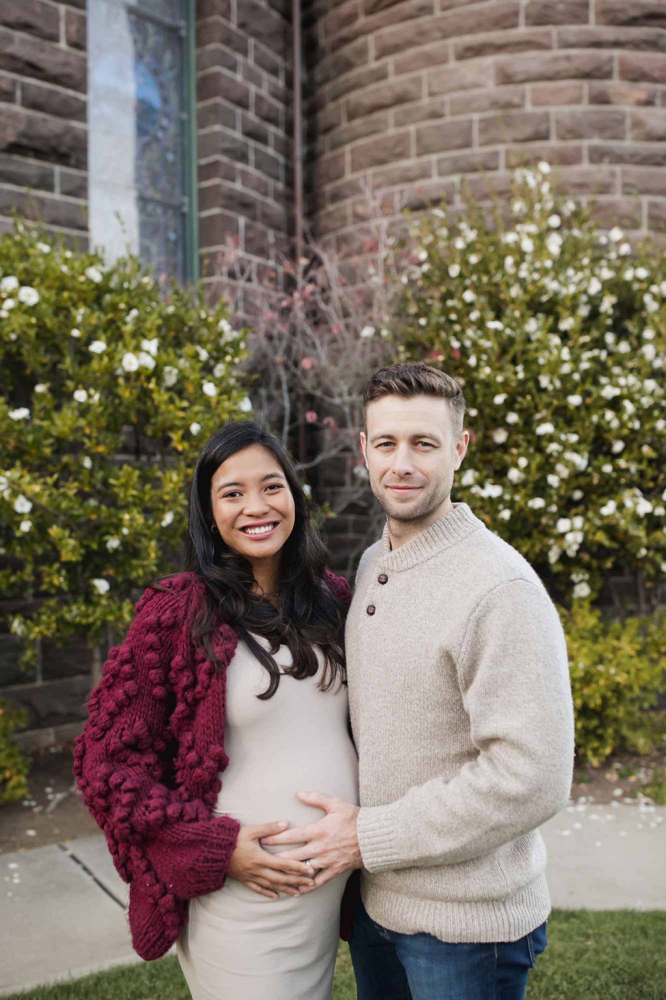 Expectant couple poses against brick building with blooming white flowers, woman in cream dress with burgundy cardigan cradling her belly