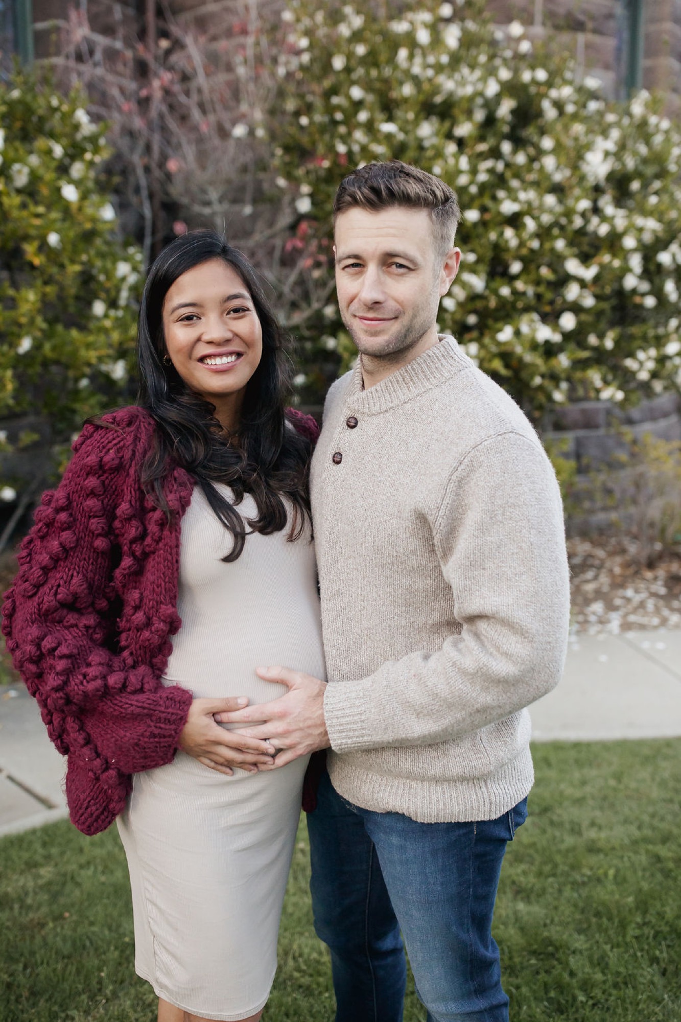 Expecting couple poses warmly together against flowering vines, with the man's hand cradling the woman's pregnant belly — San Anselmo Family Photographer